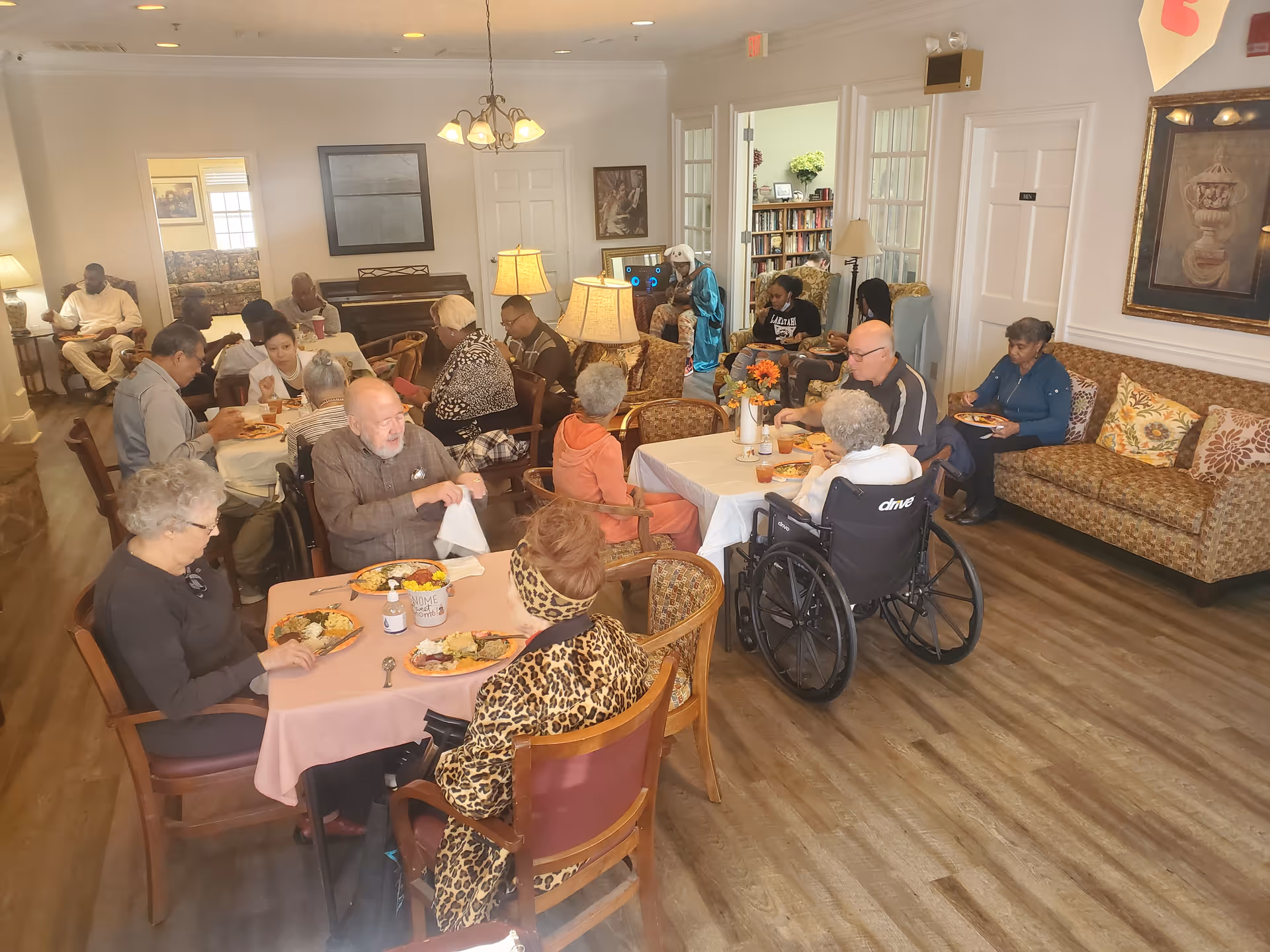 A group of senior residents eating and socializing at tables in a communal dining/living room.