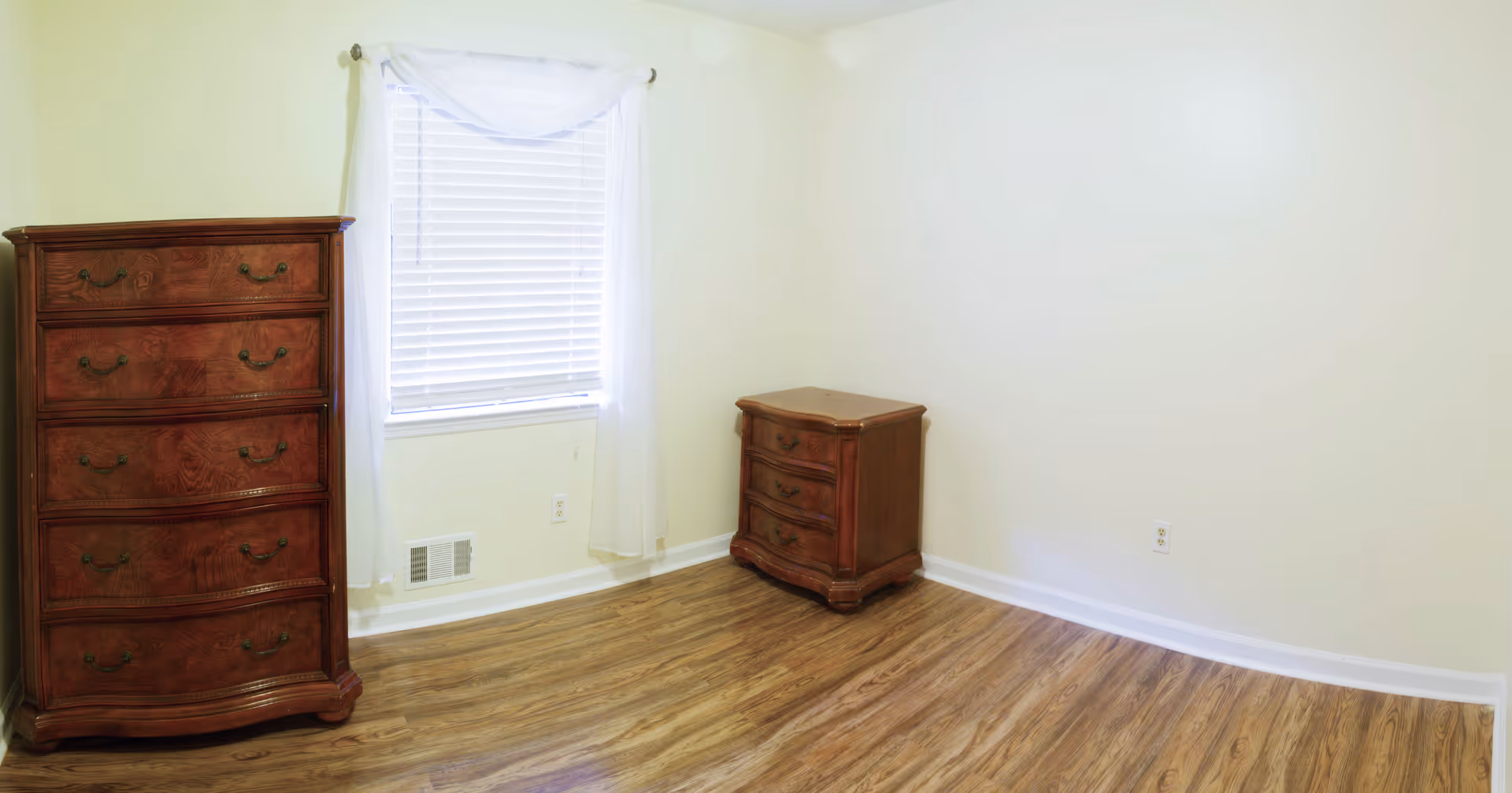 Empty room with wooden flooring, cream-colored walls, a window with white blinds and sheer curtains, a tall wooden dresser on the left, and a smaller wooden chest of drawers on the right.