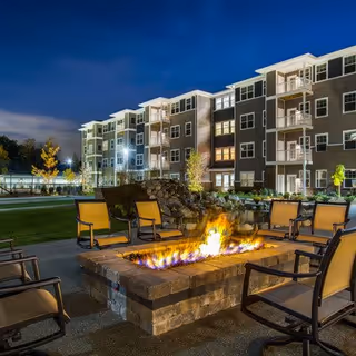 Outdoor seating area at night with a rectangular fire pit surrounded by cushioned chairs. In the background, a multi-story apartment building with balconies is illuminated, and some trees and landscaping are visible.