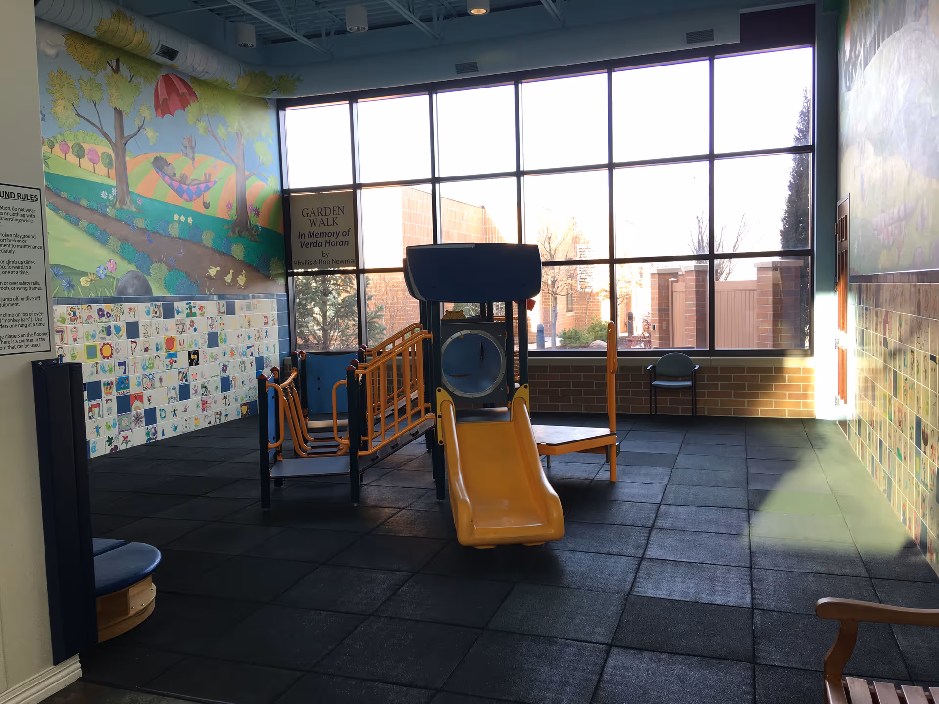 Indoor children's play area with a small yellow slide and climbing structure set on rubber tiles in front of large windows and mural-covered walls.