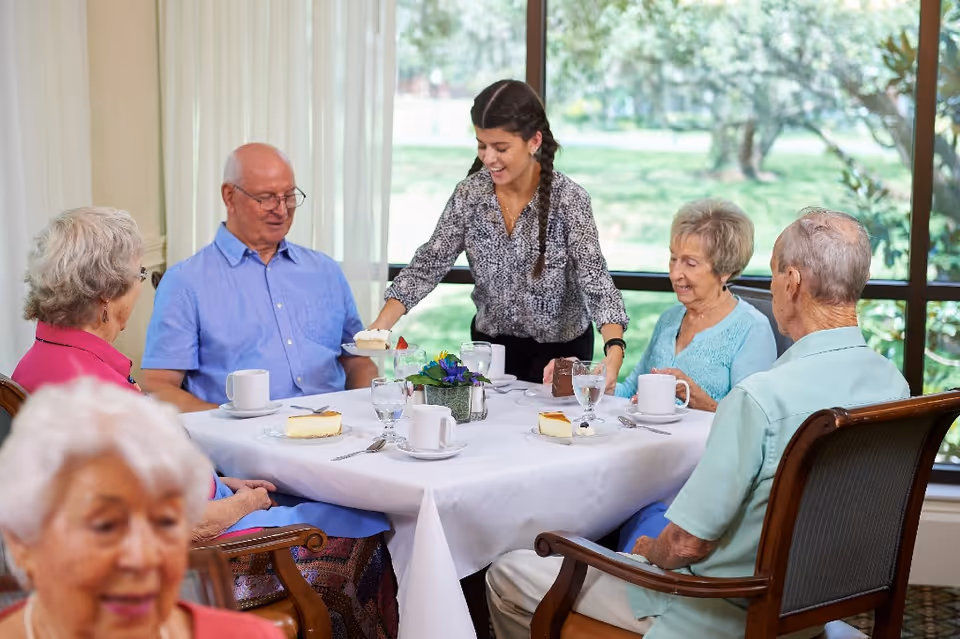 A caregiver serves dessert to a group of elderly residents seated around a dining table in a bright dining room.