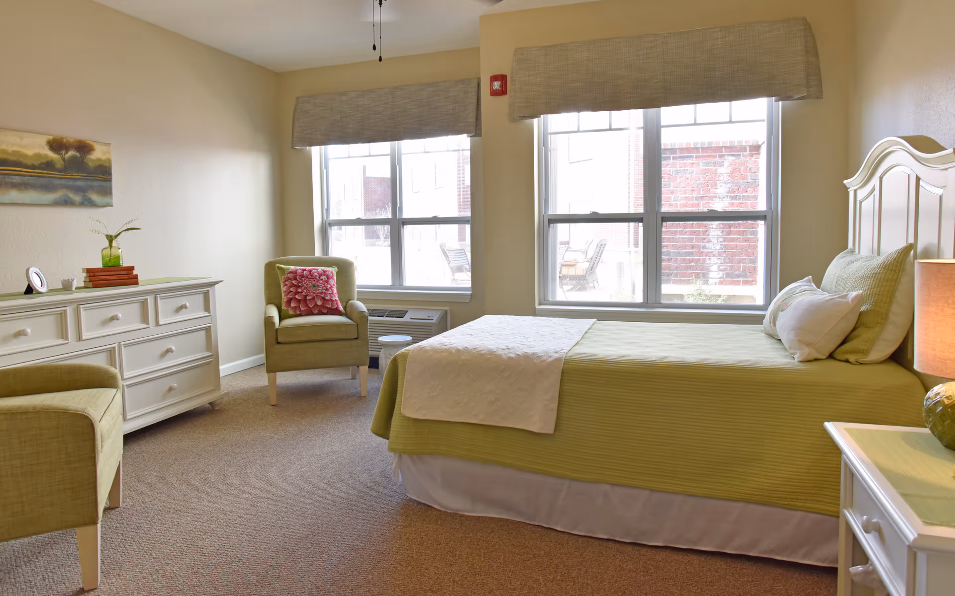 A bright and cozy bedroom in an assisted living facility featuring a bed with green bedding and white pillows, two large windows with beige valances letting in natural light, a green armchair with a pink floral cushion, a white dresser with decorative items, and a bedside table with a lamp.