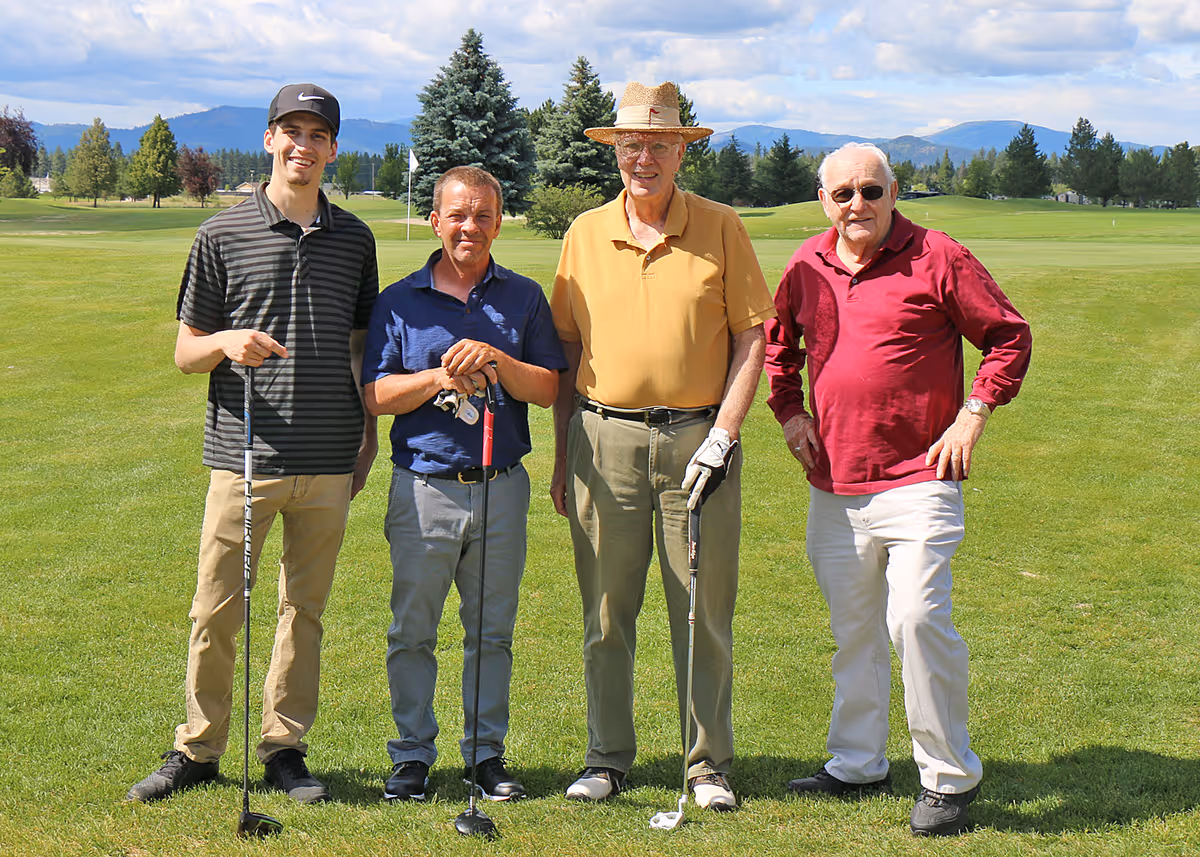 Four men standing on a golf course holding golf clubs, with trees and mountains in the background under a partly cloudy sky.