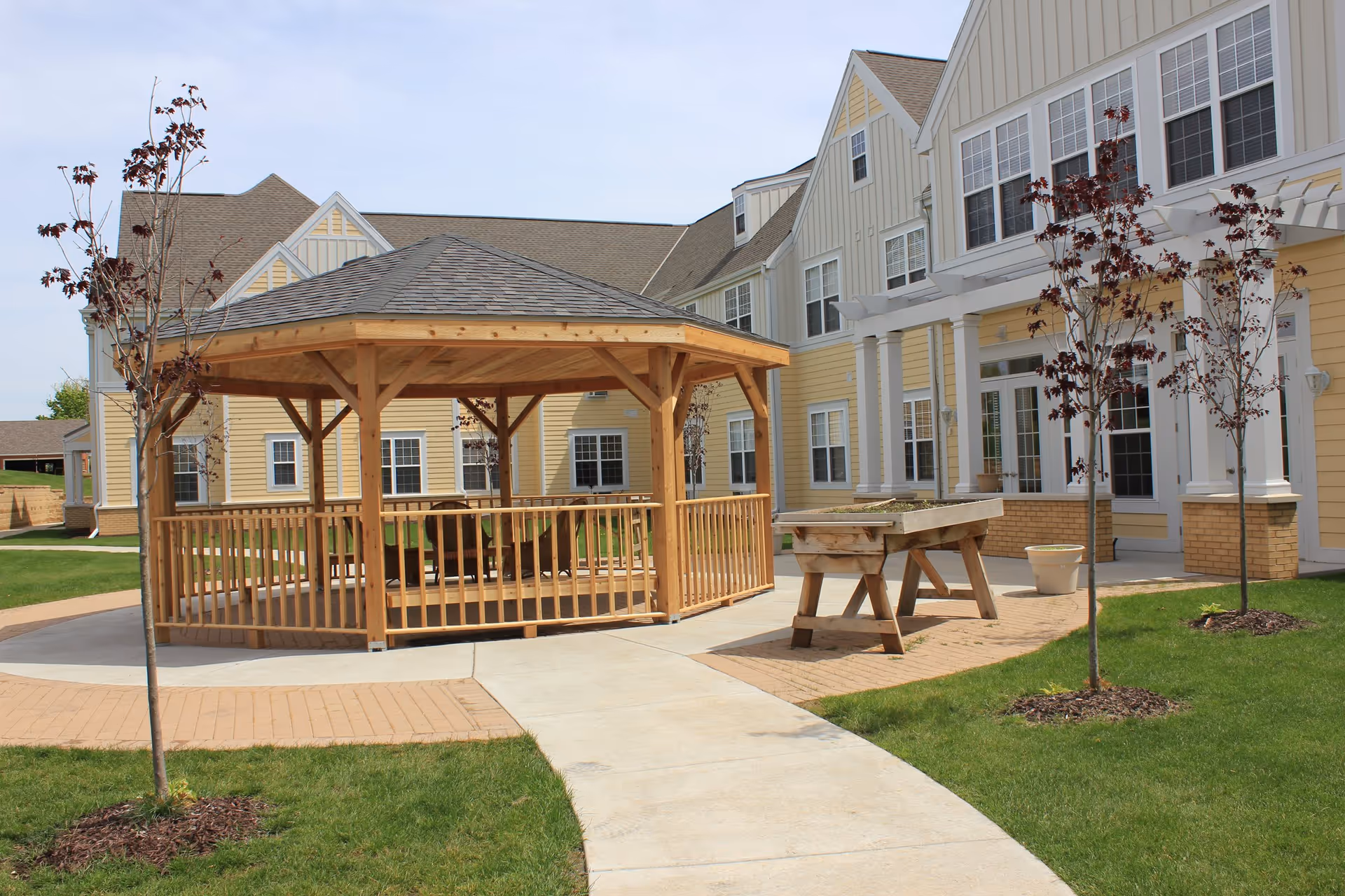 Outdoor area of a senior living facility with a wooden gazebo, a raised garden bed, young trees, and a yellow building with white trim in the background.