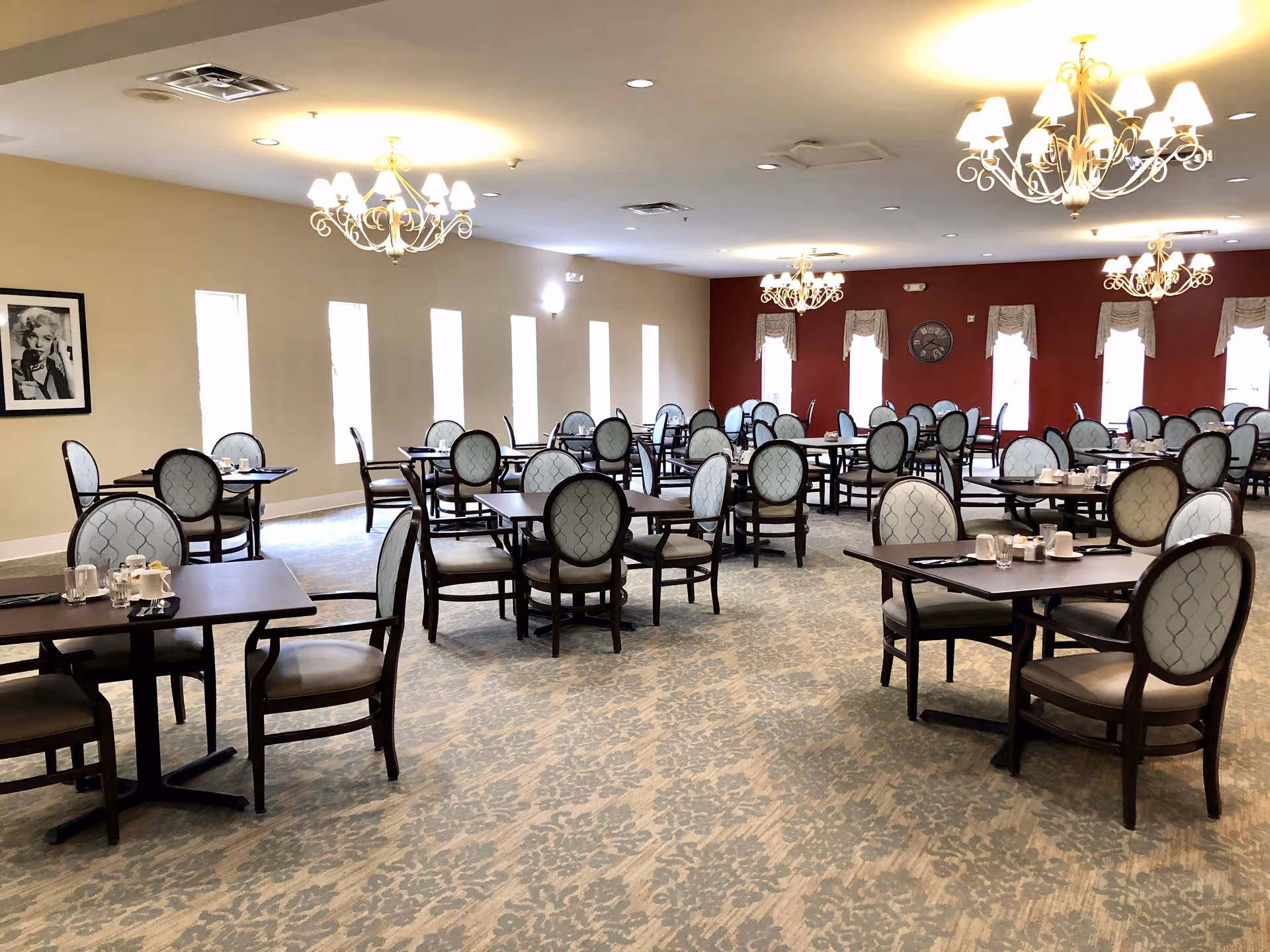 A spacious dining room with multiple tables and chairs arranged neatly. The chairs have light blue upholstery with a patterned design. The room features several chandeliers hanging from the ceiling, beige walls with a red accent wall at the far end, and multiple tall windows with curtains. A framed black and white photo is visible on one wall.
