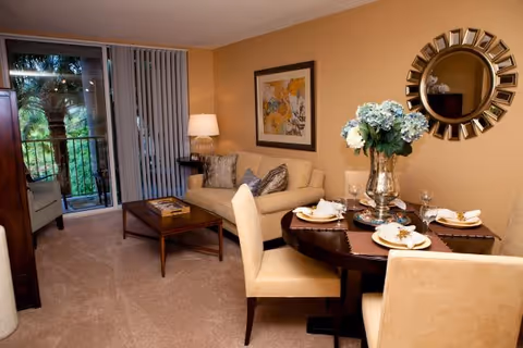 A cozy living and dining area in a senior living facility with a beige sofa, a wooden coffee table, a round dining table set for four with plates and napkins, a decorative vase with flowers, a round mirror on the wall, and a sliding glass door leading to a balcony with greenery outside.
