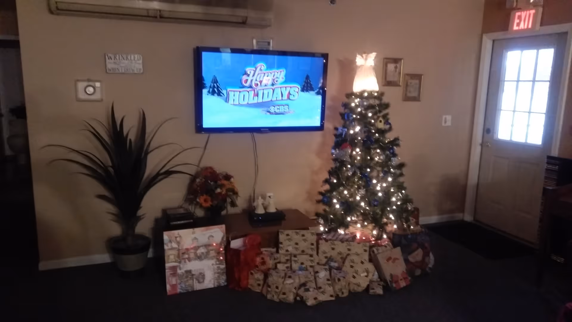 A cozy living room area decorated for the holidays with a lit Christmas tree adorned with an angel on top and surrounded by wrapped presents. A flat-screen TV mounted on the wall displays a 'Happy Holidays' message. There is a potted plant and a small table with flowers and other items beneath the TV. An exit door with a lit exit sign is visible on the right side.