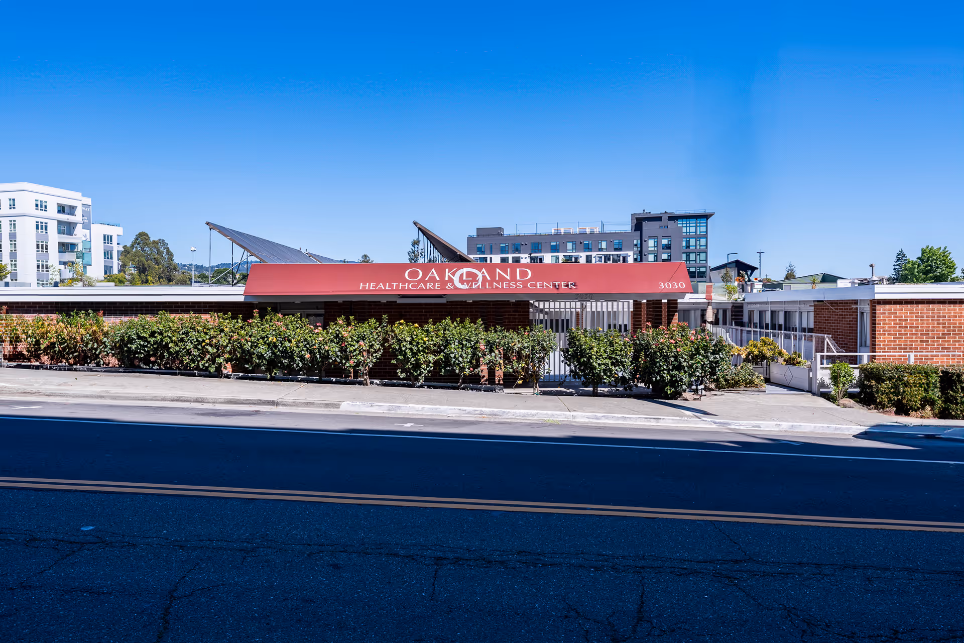 Front exterior of Oakland Healthcare & Wellness Center with a red awning, low brick wall and shrubs along a street under a clear blue sky.