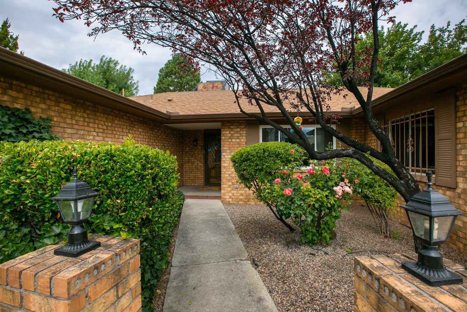 Front exterior view of a brick building with a concrete walkway leading to a door. The entrance is flanked by green bushes and flowering plants, with two black lantern-style lights mounted on brick pillars. A tree with dark red leaves is visible on the right side.