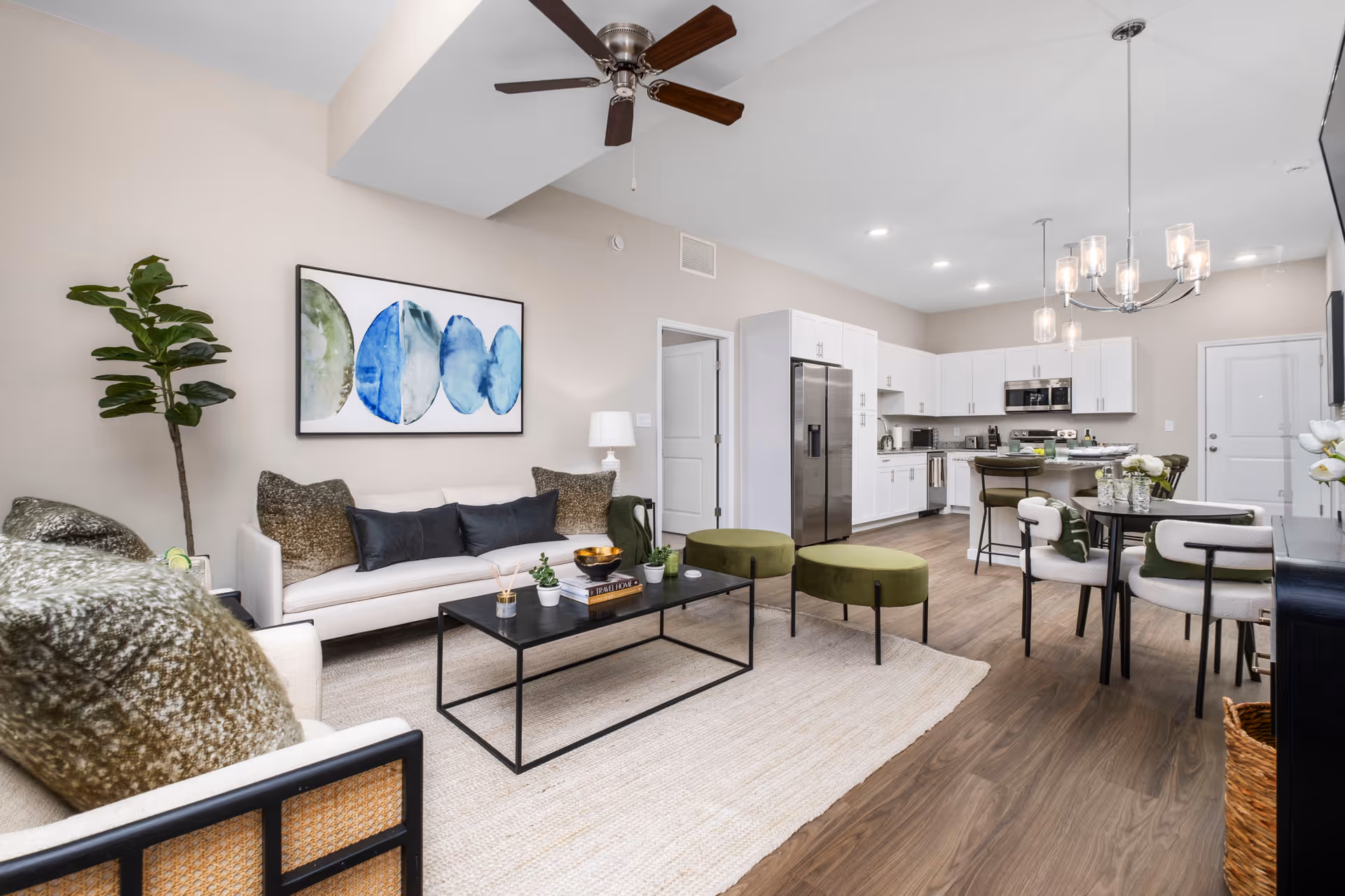A modern open-concept living room and kitchen area with light beige walls and wood flooring. The living room features a white sofa with dark and textured pillows, a black metal coffee table with small plants and decor, two green ottomans, and a large leafy plant in the corner. Above the sofa is a framed abstract artwork with blue and green circular shapes. The kitchen has white cabinets, stainless steel appliances including a refrigerator and microwave, and a kitchen island with bar stools. A dining table with four cushioned chairs is positioned near a contemporary chandelier with glass shades.