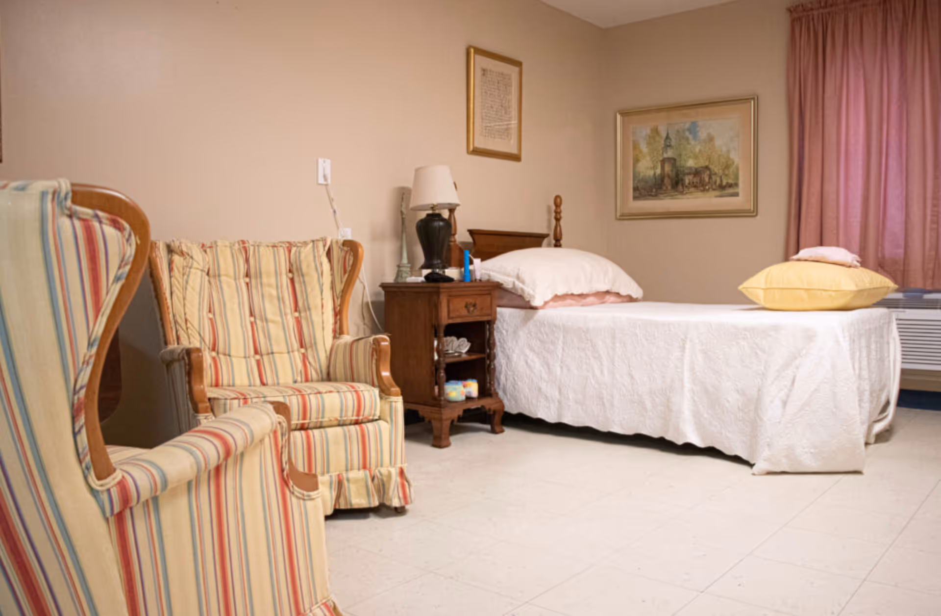 A cozy bedroom in a senior living facility featuring a single bed with white bedding and pillows, a wooden nightstand with a lamp and small items, two striped upholstered armchairs, framed artwork on the walls, and a window with pink curtains.