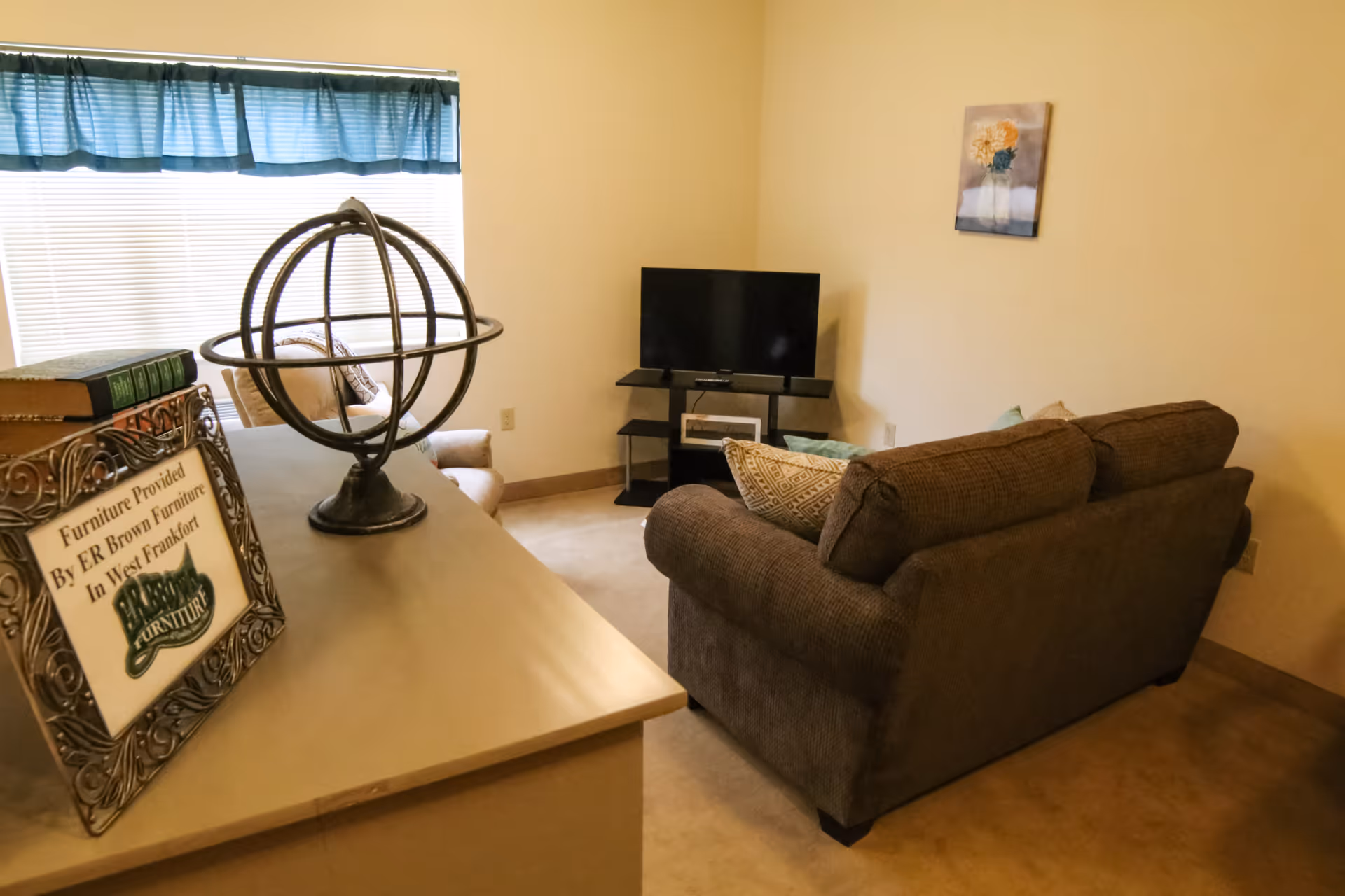 Small living room with a brown sofa facing a TV, a console table with a decorative globe and framed sign, and a window with a teal valance.