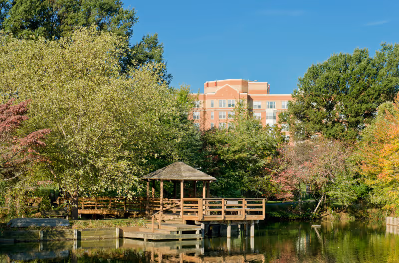 A peaceful outdoor scene at Asbury Methodist Village featuring a wooden gazebo on a dock extending over a calm pond, surrounded by lush green trees and foliage with a multi-story building visible in the background under a clear blue sky.