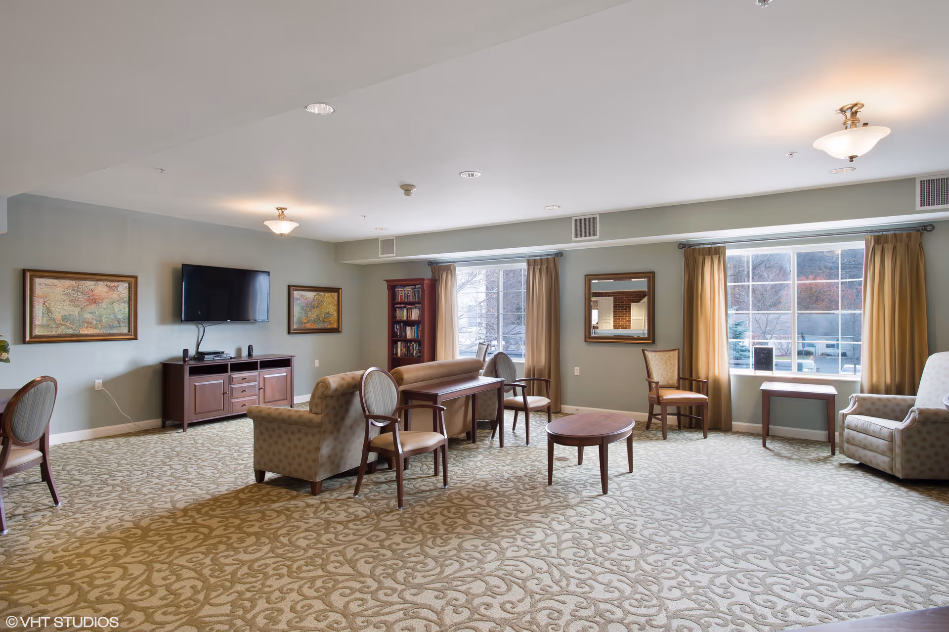 Spacious communal living room with multiple chairs and tables, a TV on a console, bookshelves, and large windows.