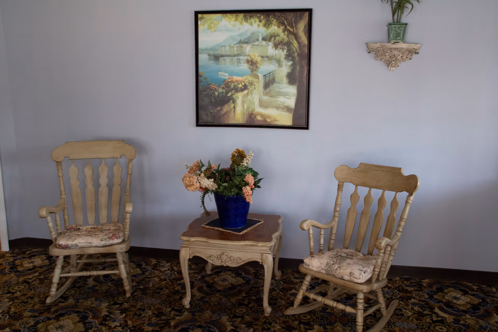 Two wooden rocking chairs with floral cushions positioned on either side of a small wooden table with a blue vase holding artificial flowers. Above the table hangs a framed painting of a scenic waterfront path with trees and flowers. A decorative wall shelf with a green plant is mounted on the wall to the right. The floor is covered with a patterned carpet.