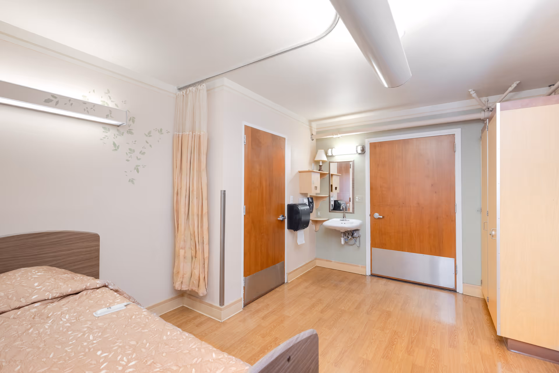 A simple room with a single bed covered in a beige patterned blanket, a privacy curtain, two wooden doors, a small wall-mounted sink with a mirror above it, a paper towel dispenser, and wooden flooring.