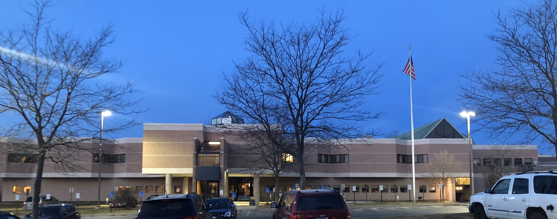 Two-story medical care facility building at dusk with a flagpole, leafless trees, and parked cars in the foreground.