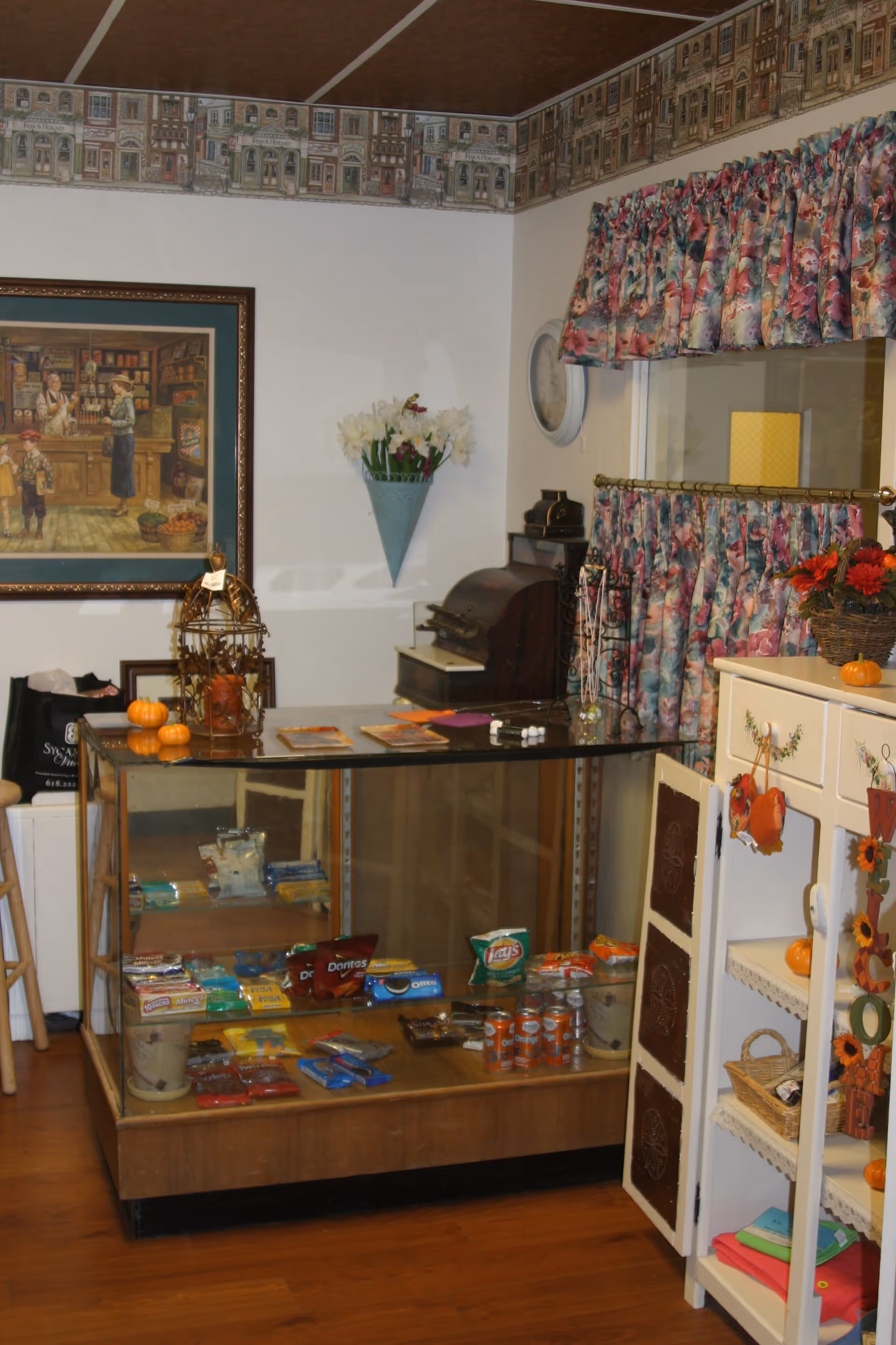 Interior view of a small shop or snack area in Sycamore Village Assisted Living, featuring a glass display case with various snacks like chips, cookies, and soda cans. There is a vintage cash register on the counter, floral curtains on the window, a framed painting on the wall, and decorative items including small pumpkins and flowers.
