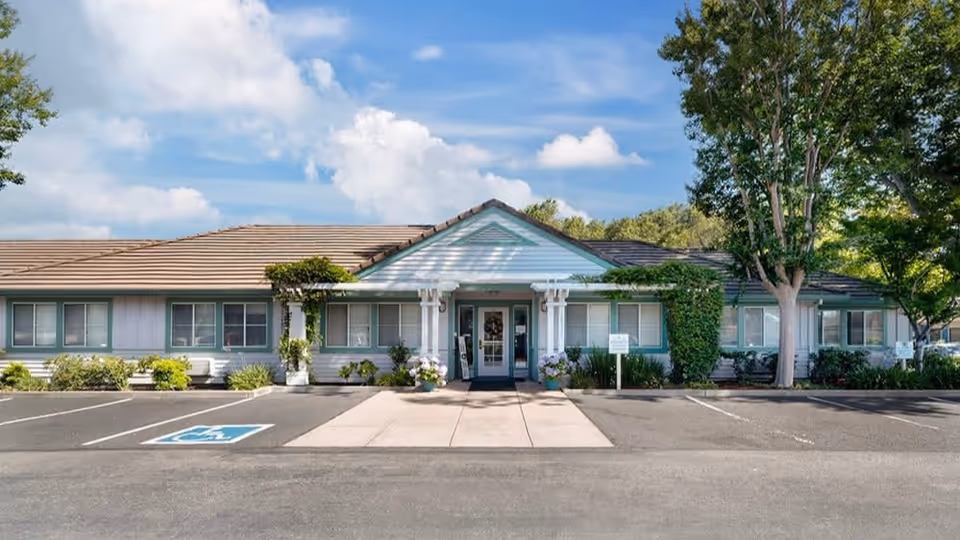 Front exterior view of a single-story building with a tiled roof, light blue siding, and green window trim. The entrance has a small covered porch with white columns and plants around it. There is a parking lot in front with a designated handicapped parking space and trees on either side of the building.
