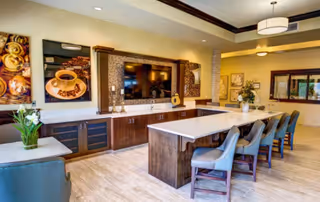 A modern communal dining and lounge area featuring a long island counter with six cushioned chairs, a large flat-screen TV mounted on a wall with decorative panels, and artwork depicting coffee cups. The room has warm lighting, wood cabinetry, and a light-colored floor.