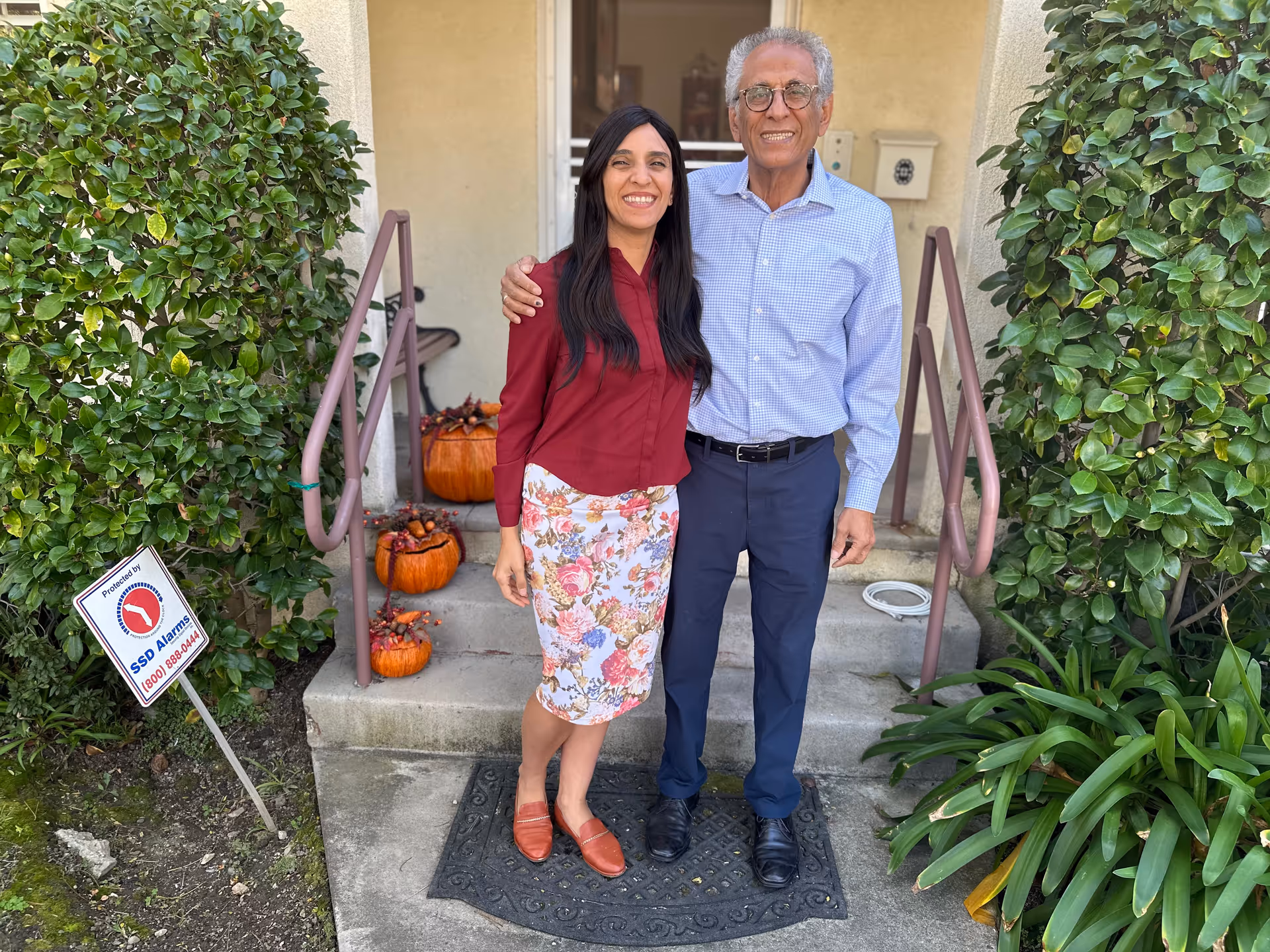 A man and a woman standing together outside the entrance of a building, smiling at the camera. The entrance has steps decorated with pumpkins and autumn-themed decorations. There are green bushes on either side of the steps and a security alarm sign on the left side.