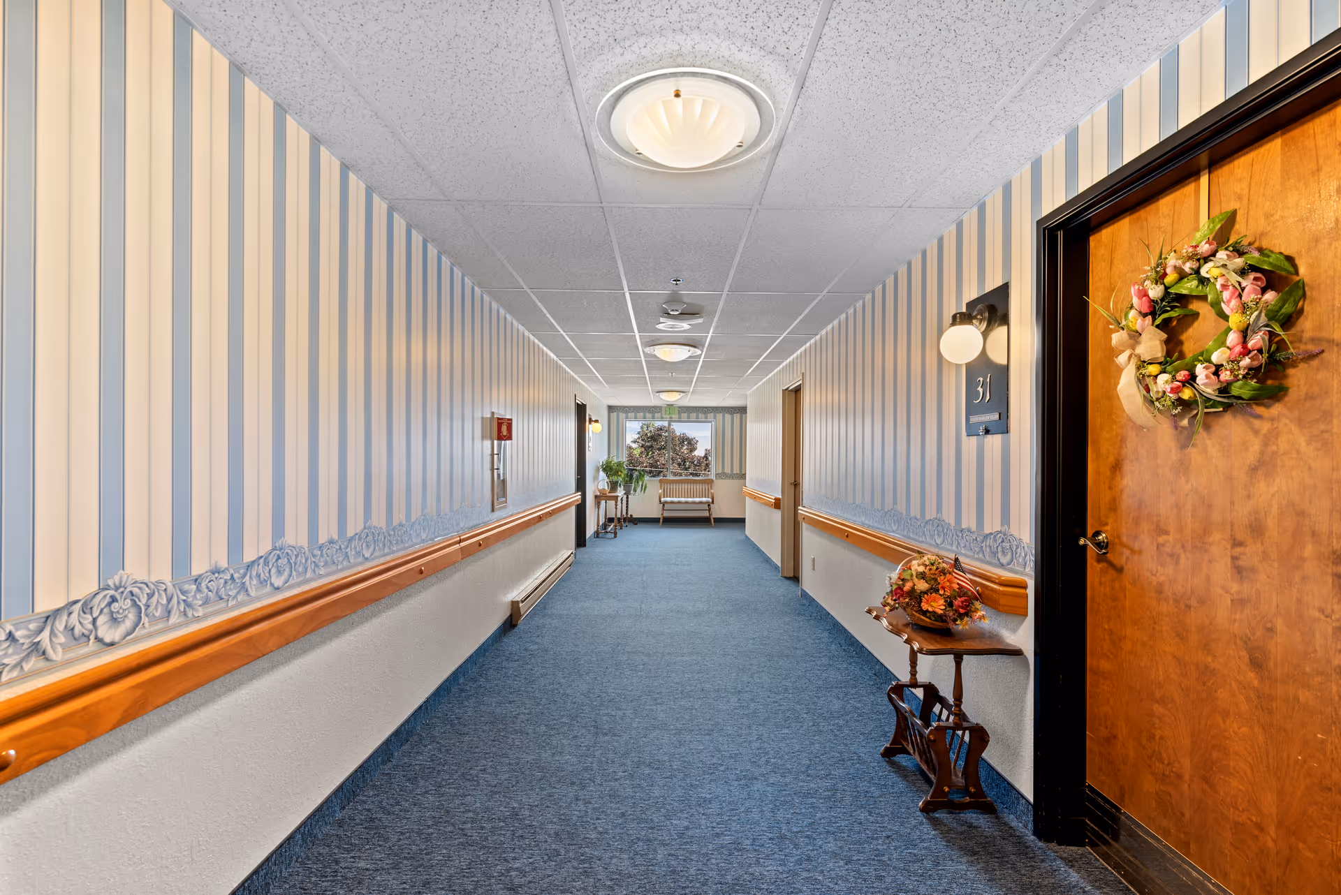 A long indoor hallway in a senior living facility with blue carpet and striped wallpaper. There is a wooden door on the right decorated with a floral wreath and a small table with a flower arrangement next to it. The hallway has ceiling lights and a window at the far end with a bench and plants nearby.