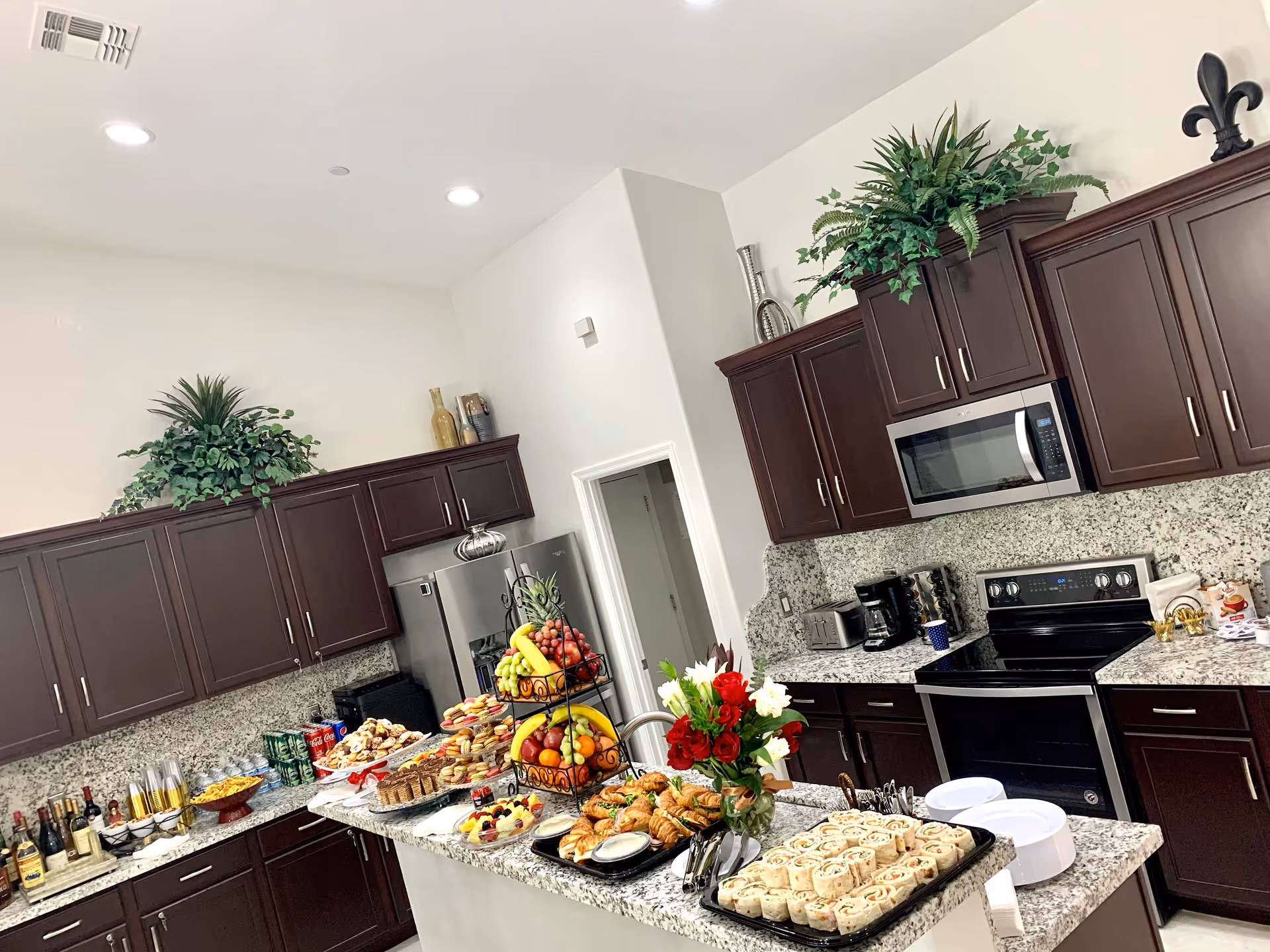 A modern kitchen with dark wood cabinets and granite countertops, featuring a variety of food items including pastries, fruit, and beverages arranged on the counter and island. There are decorative plants on top of the cabinets and a stainless steel microwave and oven.