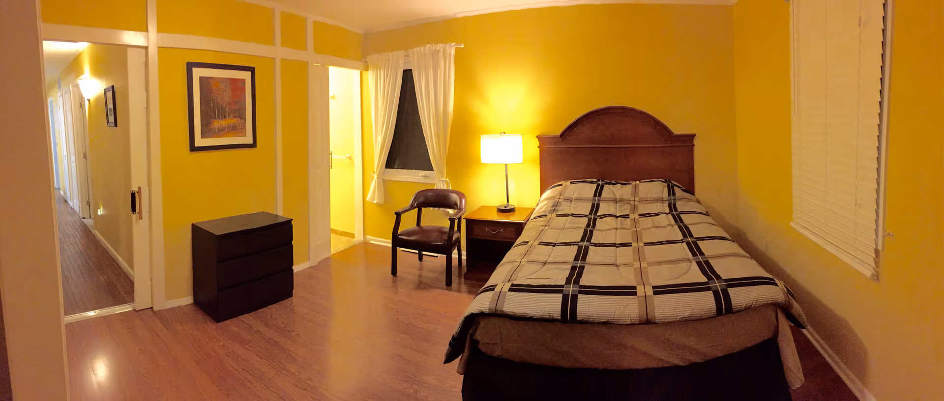 A cozy bedroom with yellow walls, a single bed with a checkered blanket, a wooden headboard, a nightstand with a lamp, a chair, a small dresser, a window with white curtains, and a hallway leading to other rooms.