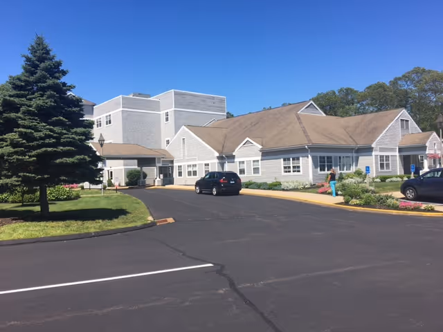 Exterior view of Bride Brook Rehabilitation and Nursing Center showing a large building with a beige roof and light gray siding. There is a paved driveway with two parked cars, a large green tree on the left, and a person walking near the entrance. The sky is clear and blue.