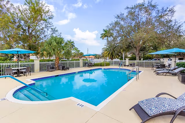 Outdoor swimming pool surrounded by lounge chairs, tables with umbrellas, and palm trees under a partly cloudy sky.