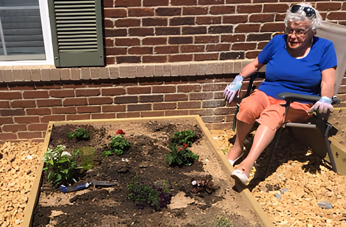 An elderly woman wearing a blue shirt, orange pants, and gardening gloves sits on a chair next to a raised garden bed with small plants and gardening tools. The scene is outdoors next to a brick wall with a window and green shutter.