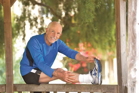An elderly man wearing a blue long-sleeve shirt and black shorts is stretching his leg while sitting on a wooden railing outdoors with greenery and flowers in the background.