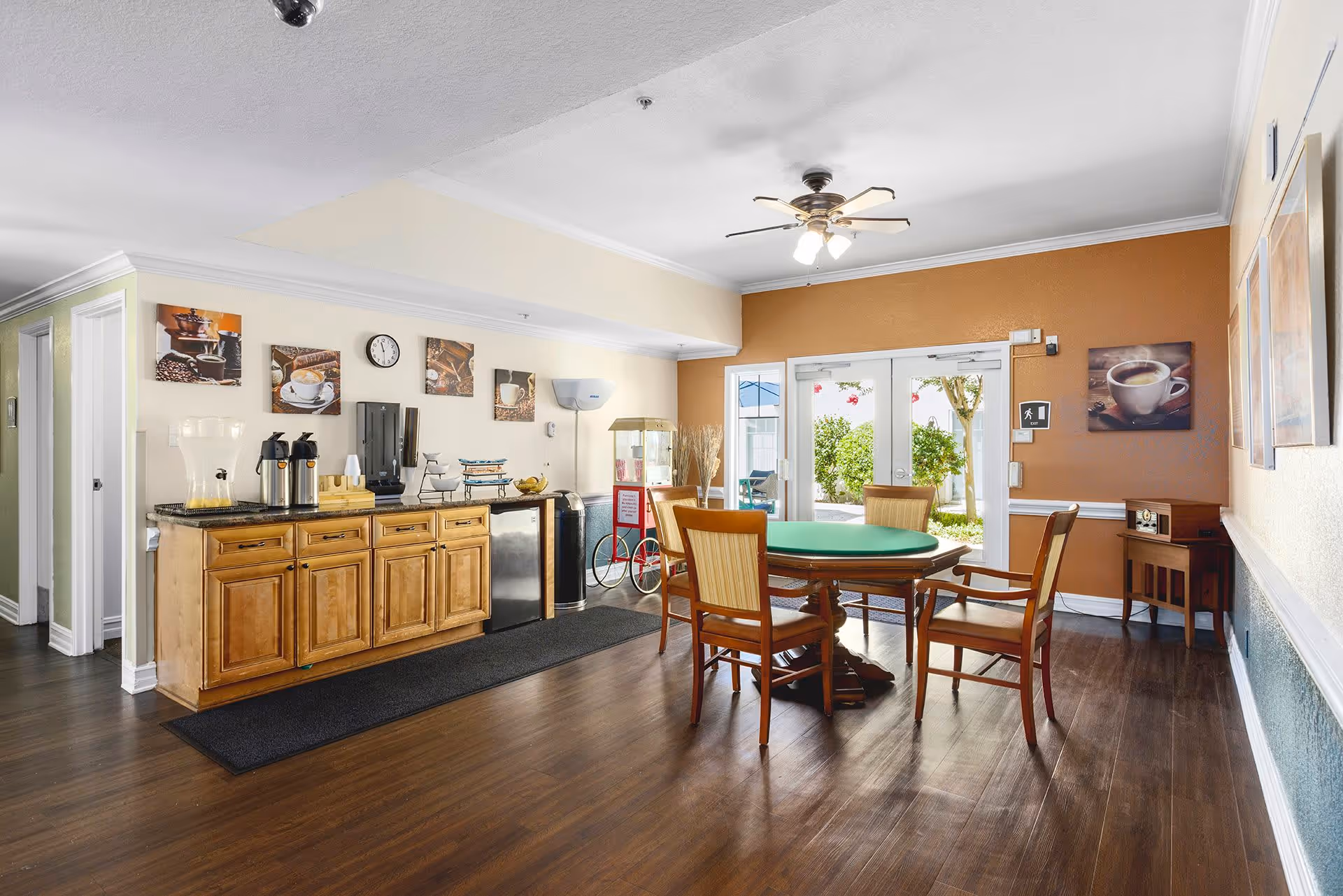 A bright common room with a round green-top table and chairs, a coffee station and cabinets along one wall, and double doors opening to an outdoor patio.