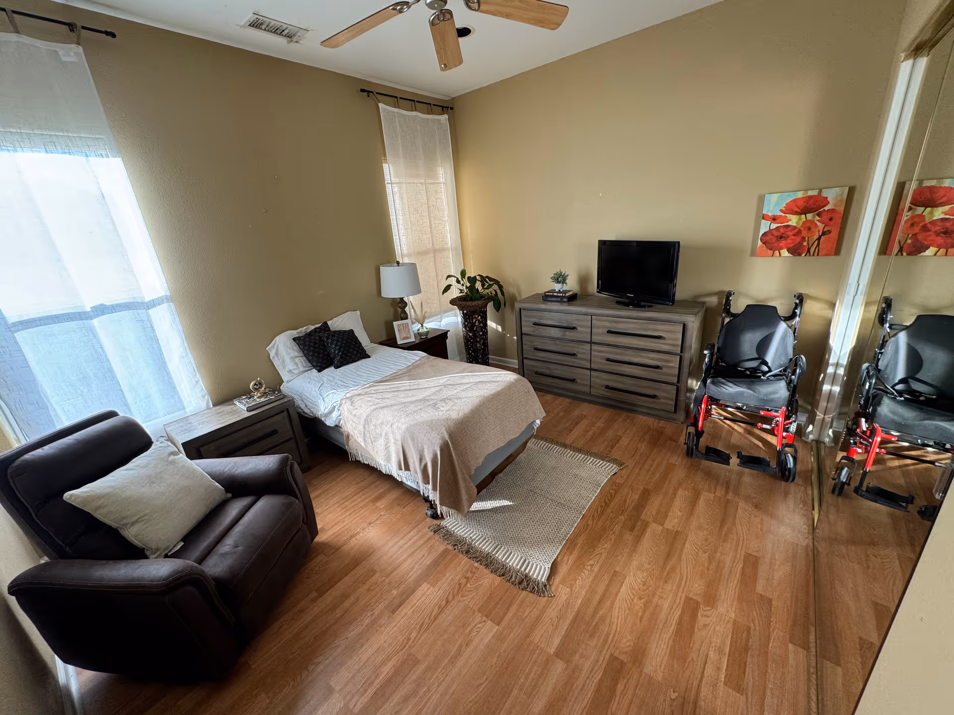 A neatly arranged bedroom with a single bed covered with white and beige bedding, a dark brown recliner chair with a light-colored pillow, a wooden nightstand with a lamp and decorative items, a wooden dresser with a small TV and books on top, a wheelchair next to the dresser, two windows with sheer curtains, a ceiling fan, and a wall painting of red flowers. The floor is wooden, and there is a beige rug under the bed.