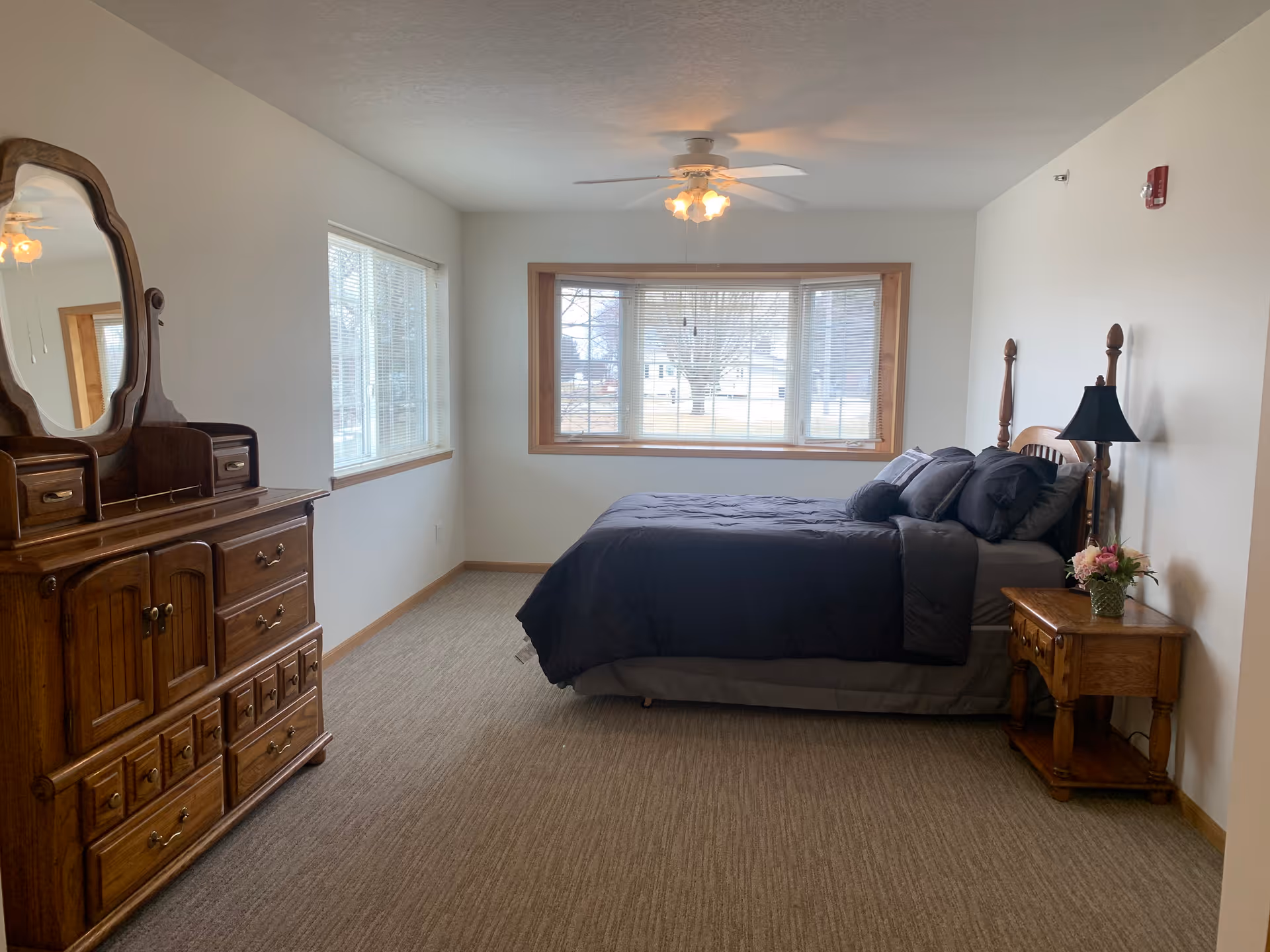 A bedroom with a large window featuring blinds, a bed with dark bedding and multiple pillows, a wooden nightstand with a lamp and a flower vase, and a wooden dresser with a mirror. The room has beige carpet and white walls with wood trim.