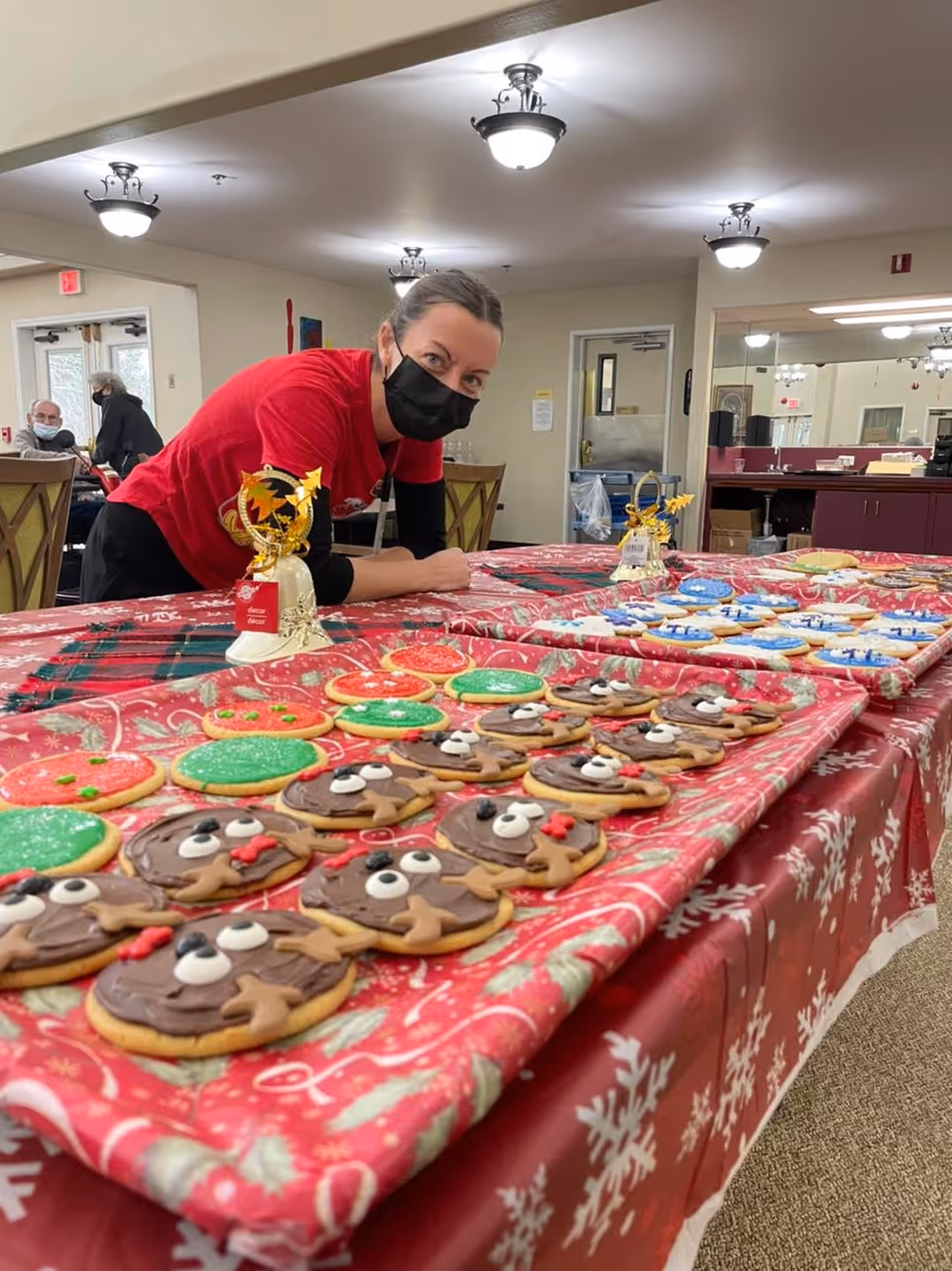 A woman wearing a black face mask and a red shirt leans over a table covered with a red holiday-themed tablecloth. The table is filled with decorated Christmas cookies shaped like reindeer, green and red circles, and other festive designs. In the background, there are chairs, a door, and a mirror reflecting part of the room.