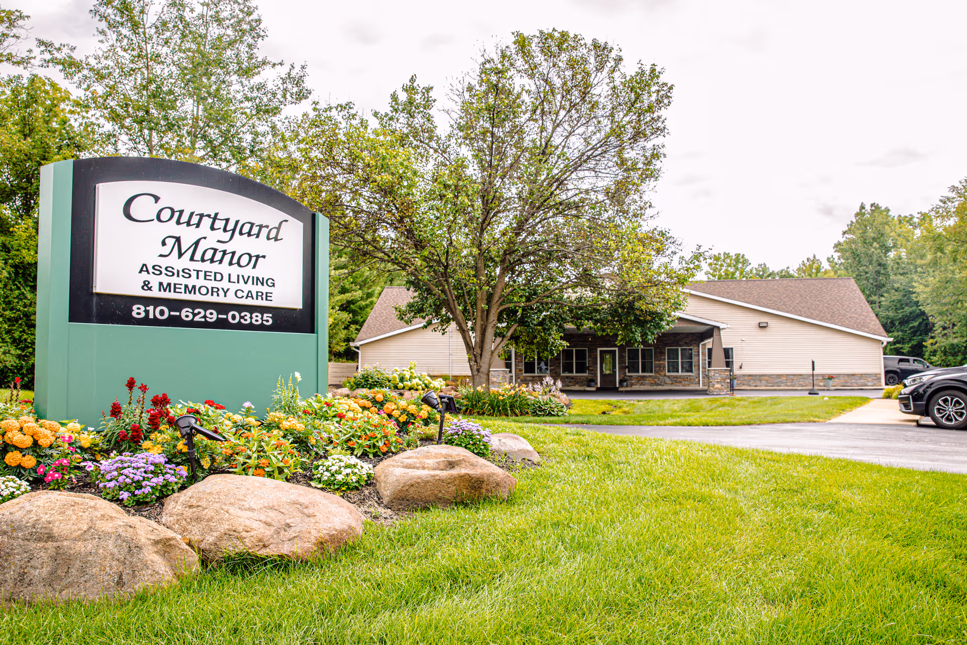 Outdoor view of Courtyard Manor assisted living and memory care facility with a large sign displaying the facility name and phone number, surrounded by colorful flowers and rocks, with a building and parked cars in the background.
