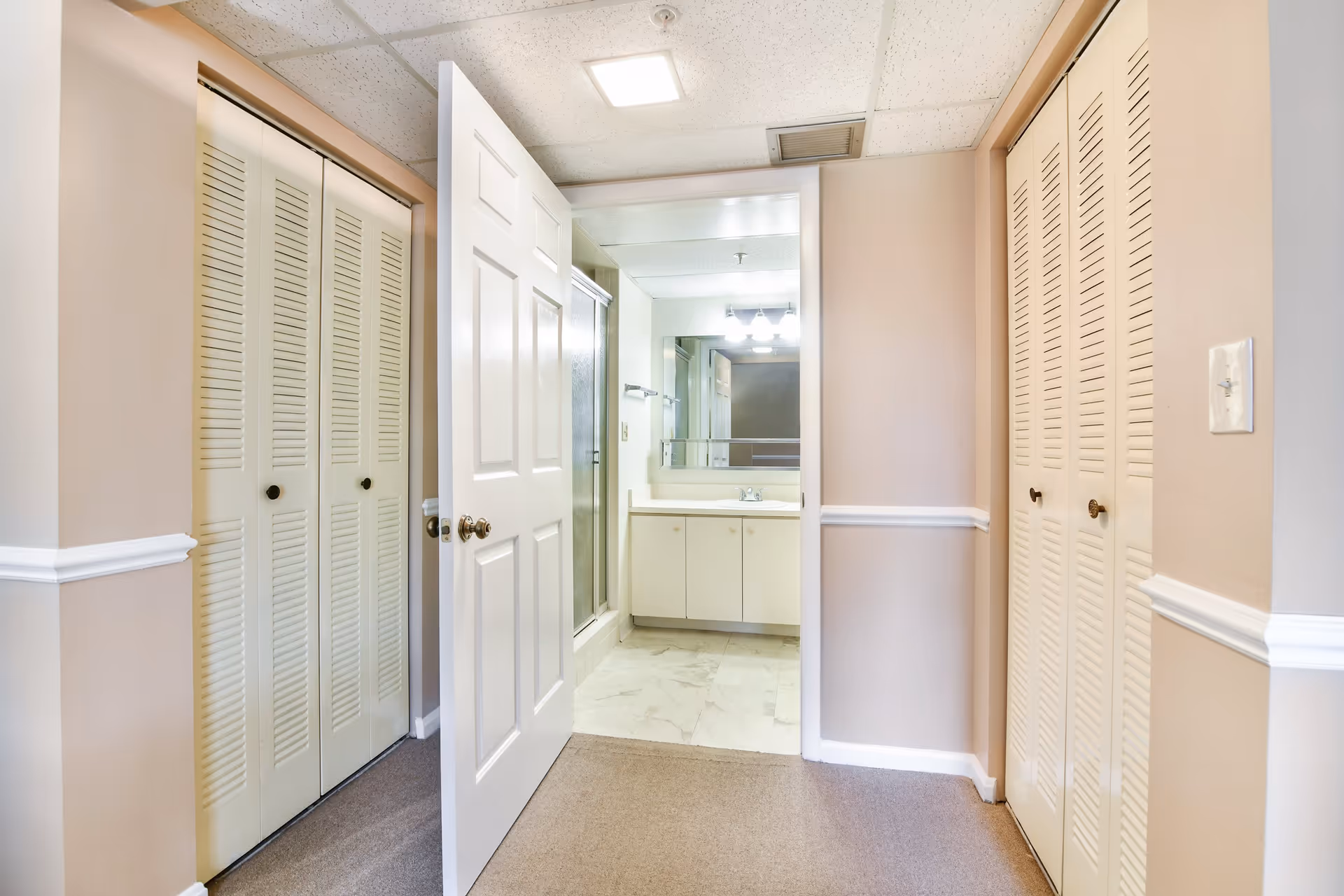 View of a hallway with beige walls and white trim leading to a bathroom. The bathroom has a large mirror, a sink with cabinets underneath, and a glass shower enclosure. There are two sets of louvered closet doors on either side of the hallway.