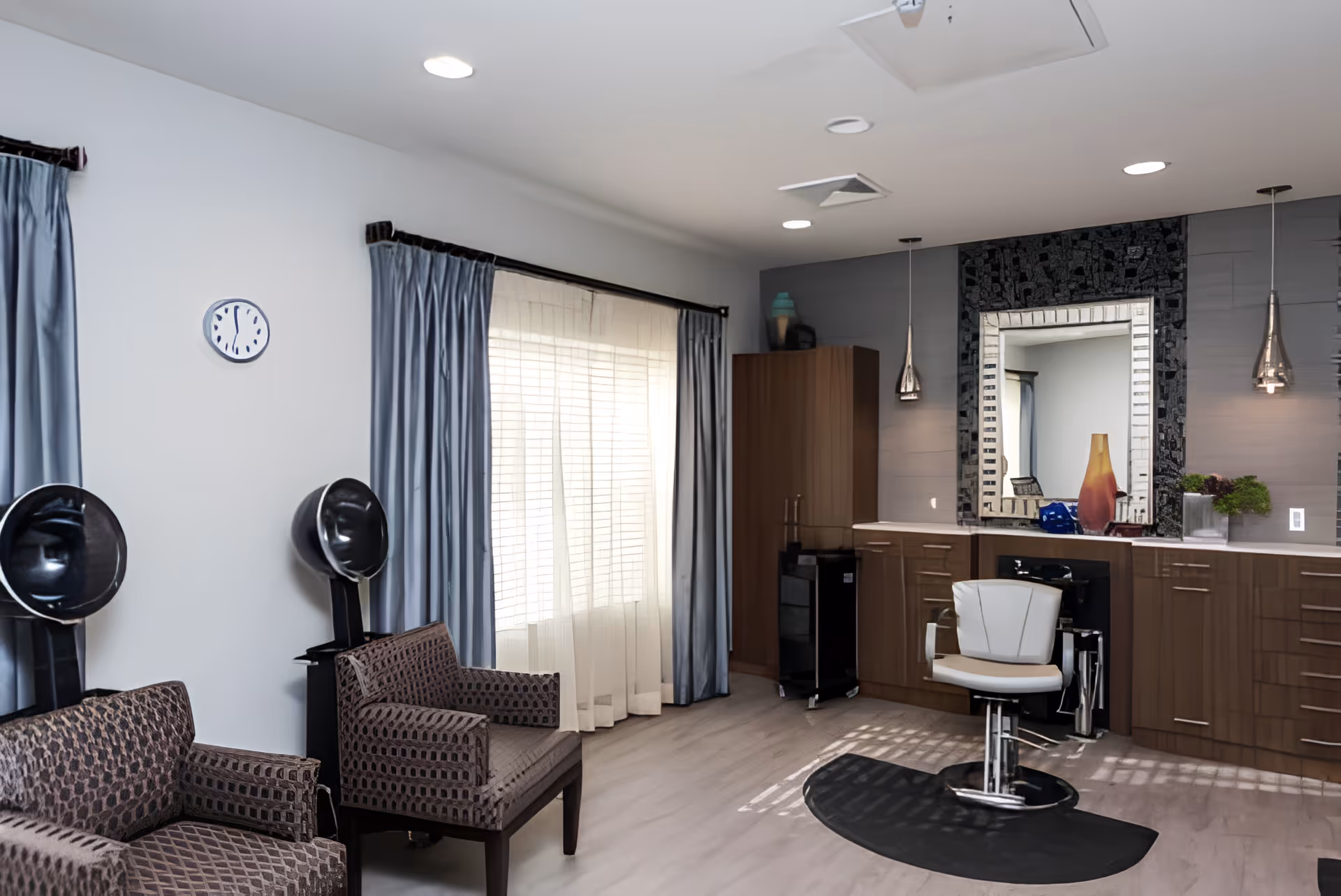 Salon-style interior with a styling chair and mirror, cabinets, seating with hooded hair dryers, and a curtained window.