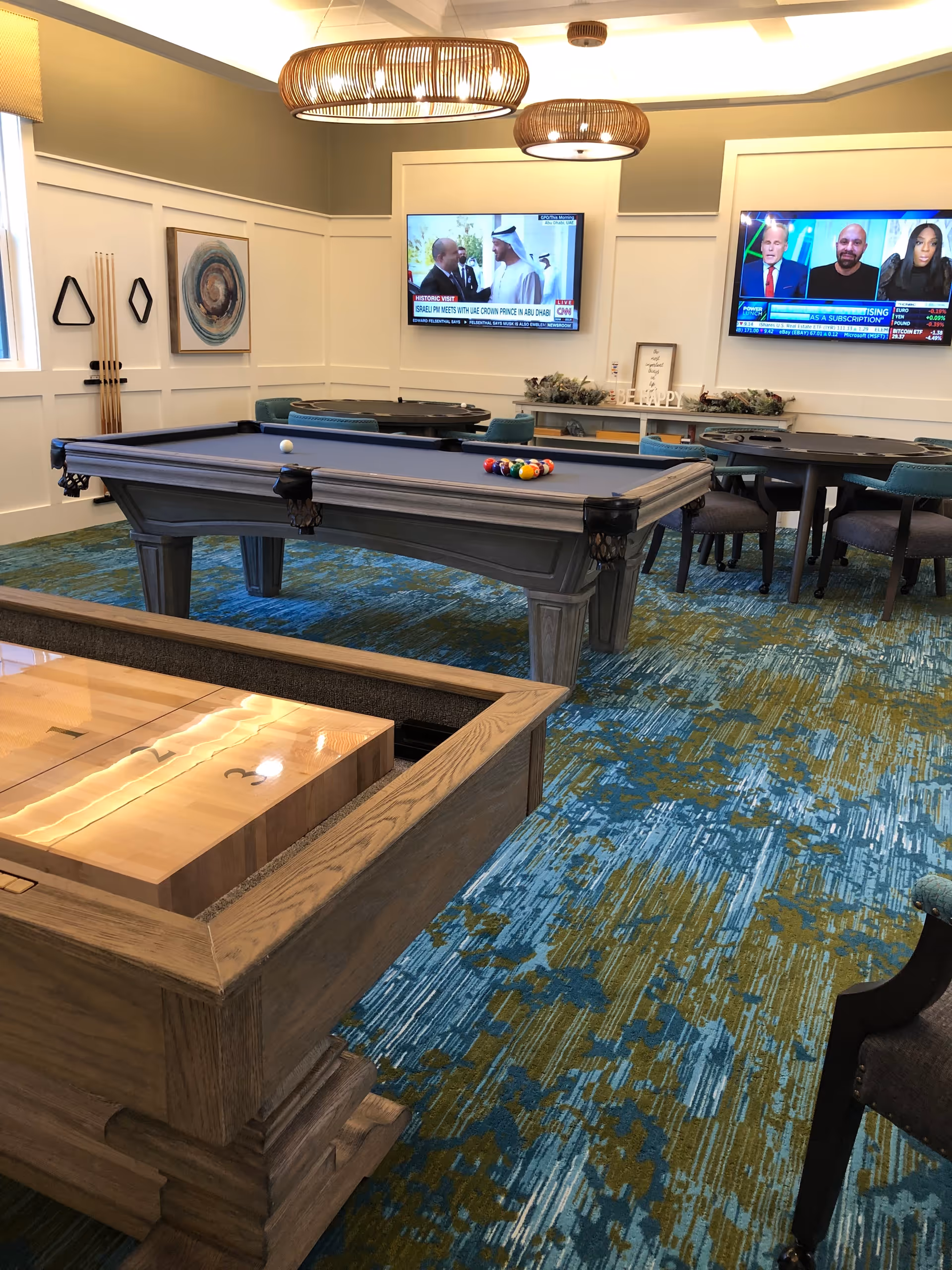 A recreational room with a pool table and a shuffleboard table on a blue and green patterned carpet. The room has two flat-screen TVs mounted on the wall, one showing a news broadcast and the other showing a talk show. There are several chairs and tables around the room, with two large wicker pendant lights hanging from the ceiling. Pool cues are mounted on the wall next to a window with a beige shade.