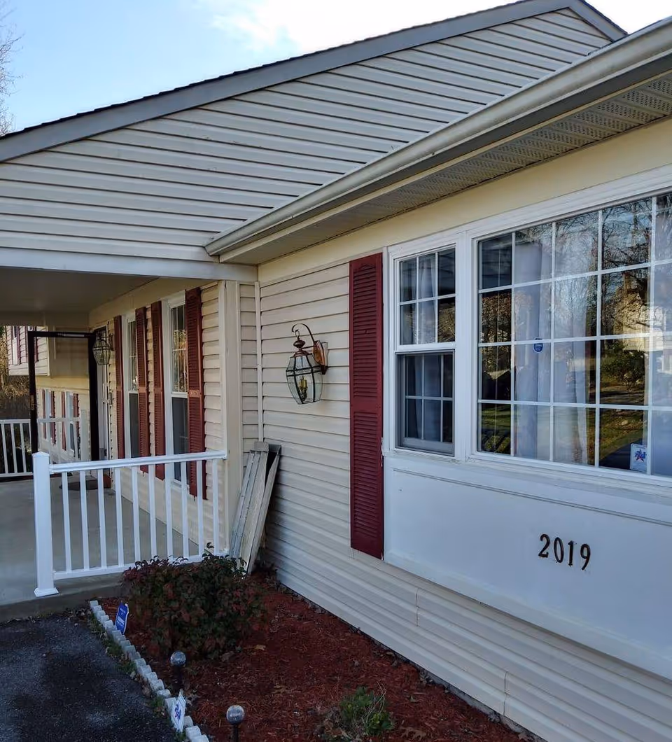 Front exterior of a beige-siding senior living facility with red shutters and a small porch.