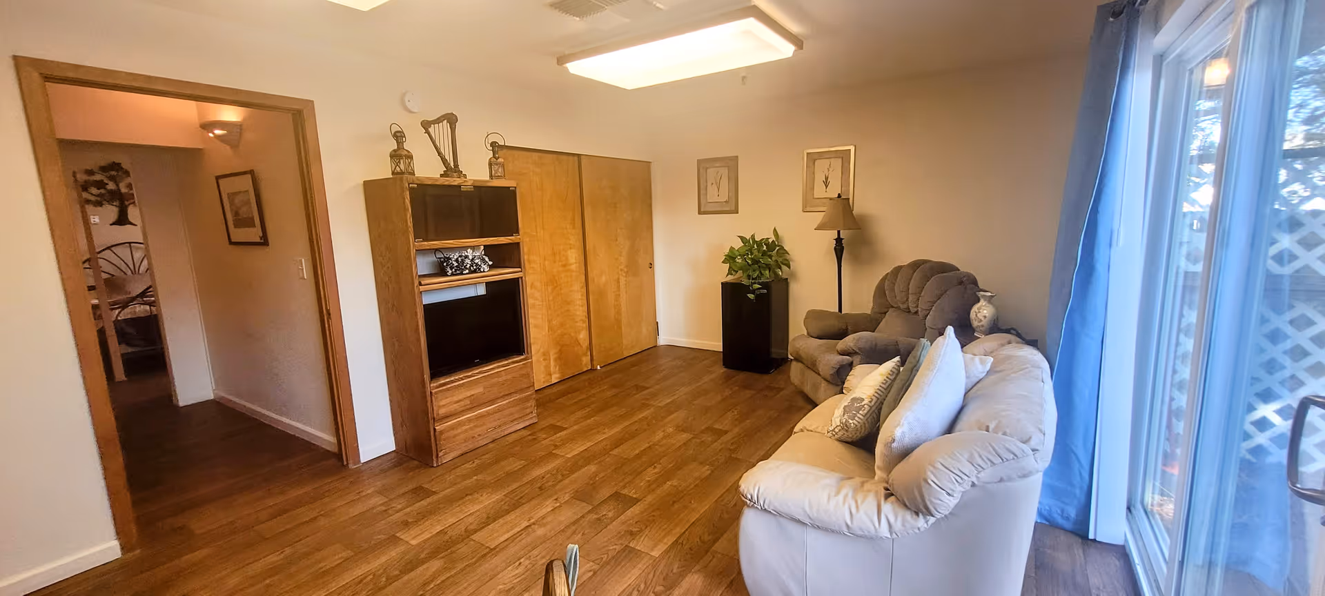A cozy living room with wooden flooring, a beige couch with pillows, a gray armchair, a tall black floor lamp, a small plant on a black stand, and a wooden entertainment center with a TV. There is a sliding glass door with blue curtains on the right side, and an open doorway leading to another room on the left.