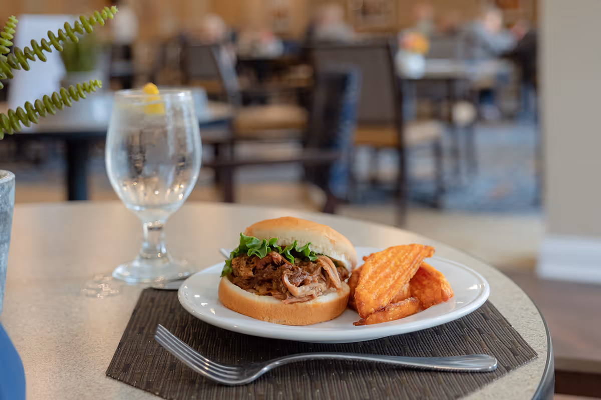 A plate with a pulled pork sandwich topped with lettuce and a side of waffle fries on a table in a dining area. Next to the plate is a glass of water with a lemon wedge. The background shows blurred tables and chairs in a communal dining room.