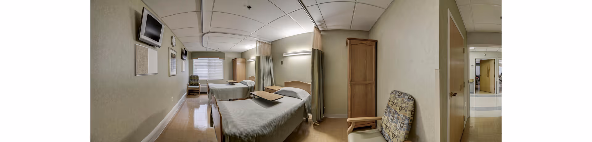 A hospital-style room with two beds separated by a curtain. Each bed has a tray table, and there is a patterned armchair near the door. The room has a wall-mounted TV, a clock, and a window with blinds. The walls are light-colored, and the floor is a polished light brown.