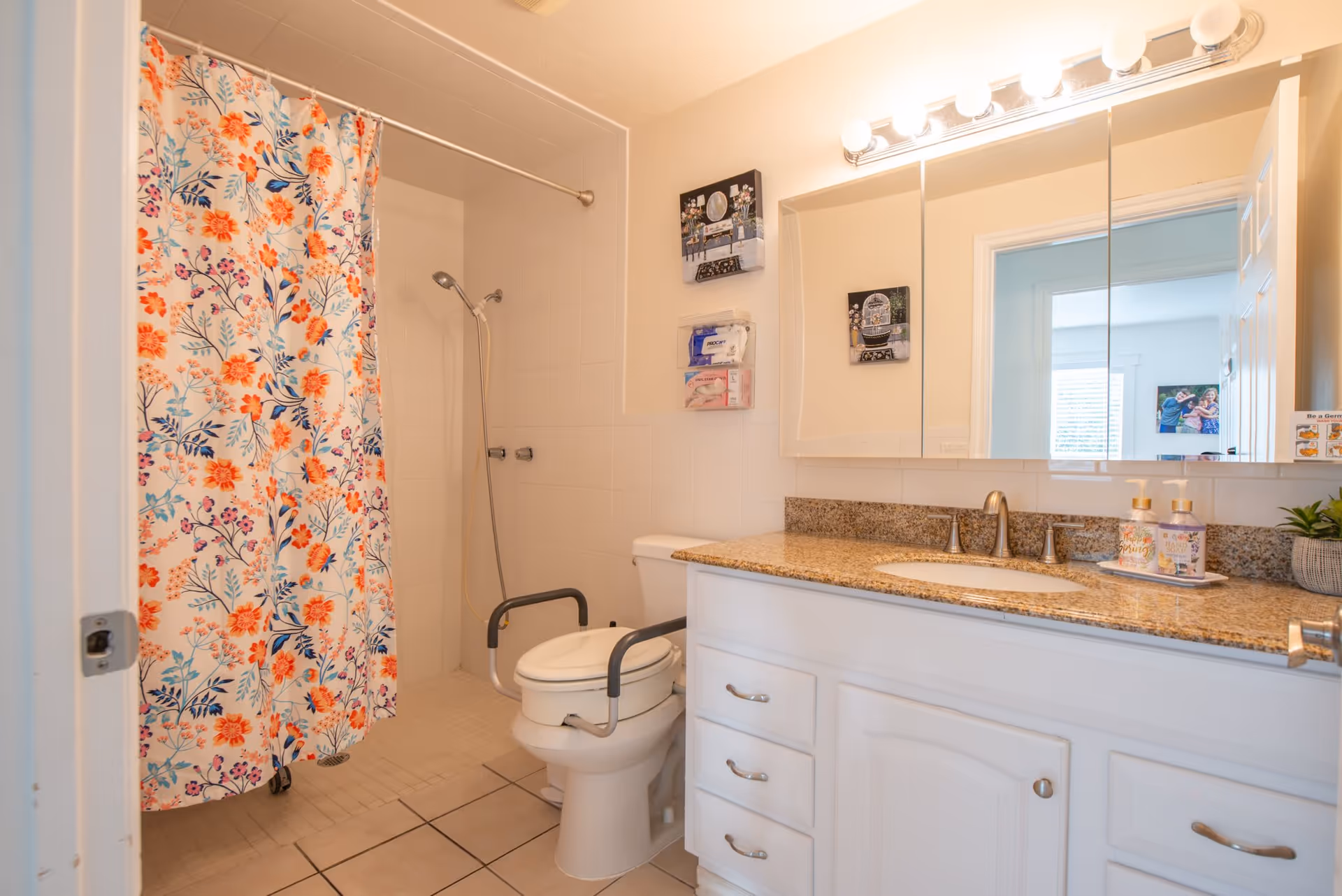 A bathroom with a white toilet equipped with safety rails, a shower area with a floral shower curtain, a granite countertop with a sink, and a large mirror above the sink. The bathroom has white tiled walls and floor, and there are two small decorative pictures on the wall.