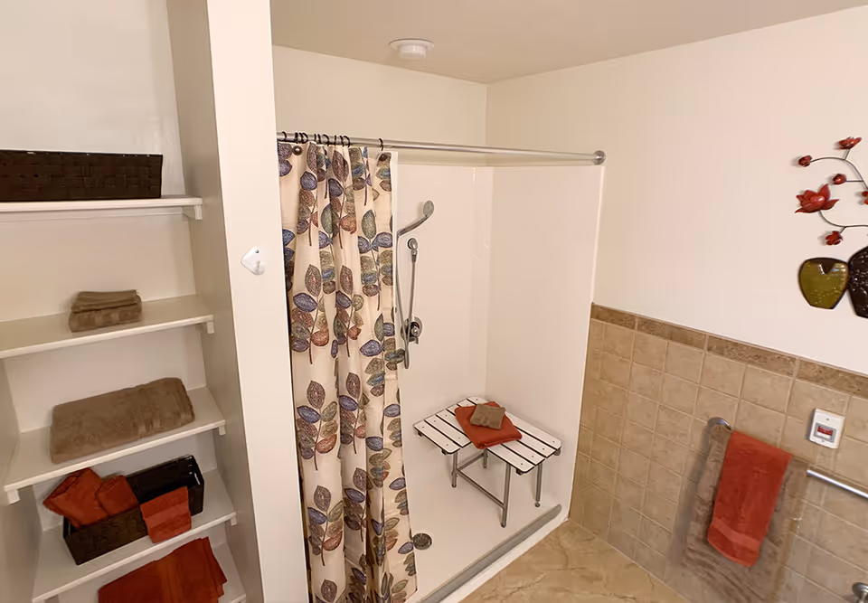 Bathroom with a walk-in shower featuring a leaf-patterned curtain, a fold-up shower bench, shelving with towels, and tiled half-wall with grab bar.