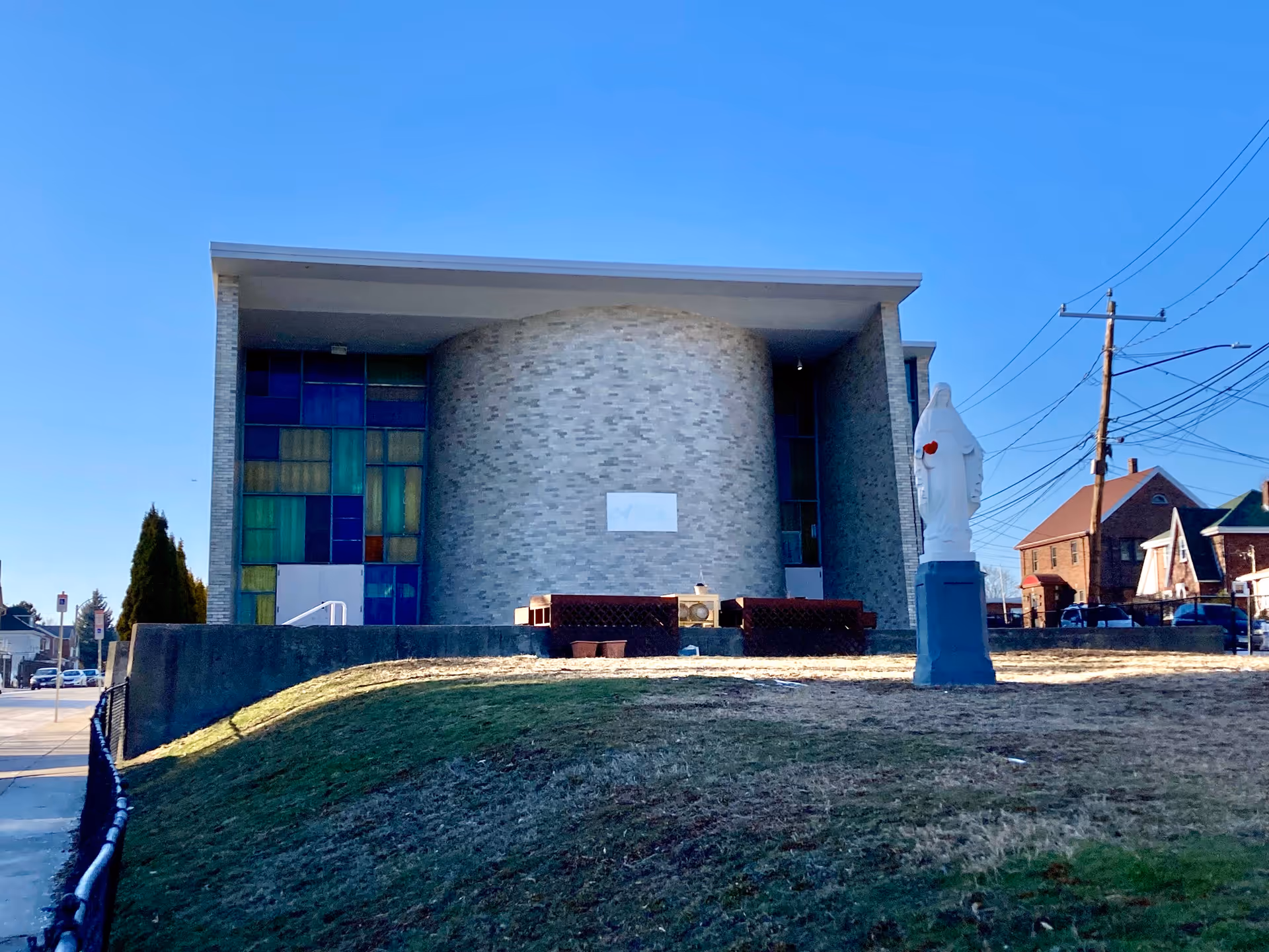 Exterior view of a modern building with a curved brick facade and colorful stained glass windows on the left side. In front of the building, there is a statue of a religious figure on a pedestal. The sky is clear and blue, and there are houses and utility poles visible in the background.