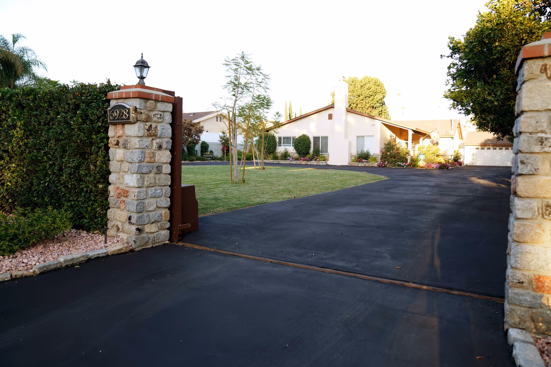 View of the entrance driveway to a residential-style senior living facility with a stone pillar displaying the address number 6928, a paved driveway leading to a single-story building surrounded by well-maintained landscaping including grass, bushes, and small trees.