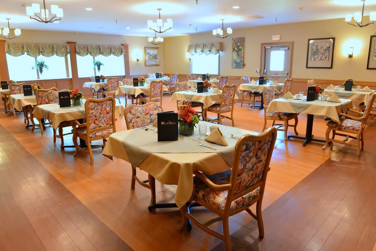 A bright and spacious dining room with multiple tables covered in beige tablecloths, each set with napkins, glasses, and floral centerpieces. The chairs have floral upholstery, and the room features wooden flooring, large windows with valances, framed artwork on the walls, and ceiling light fixtures.