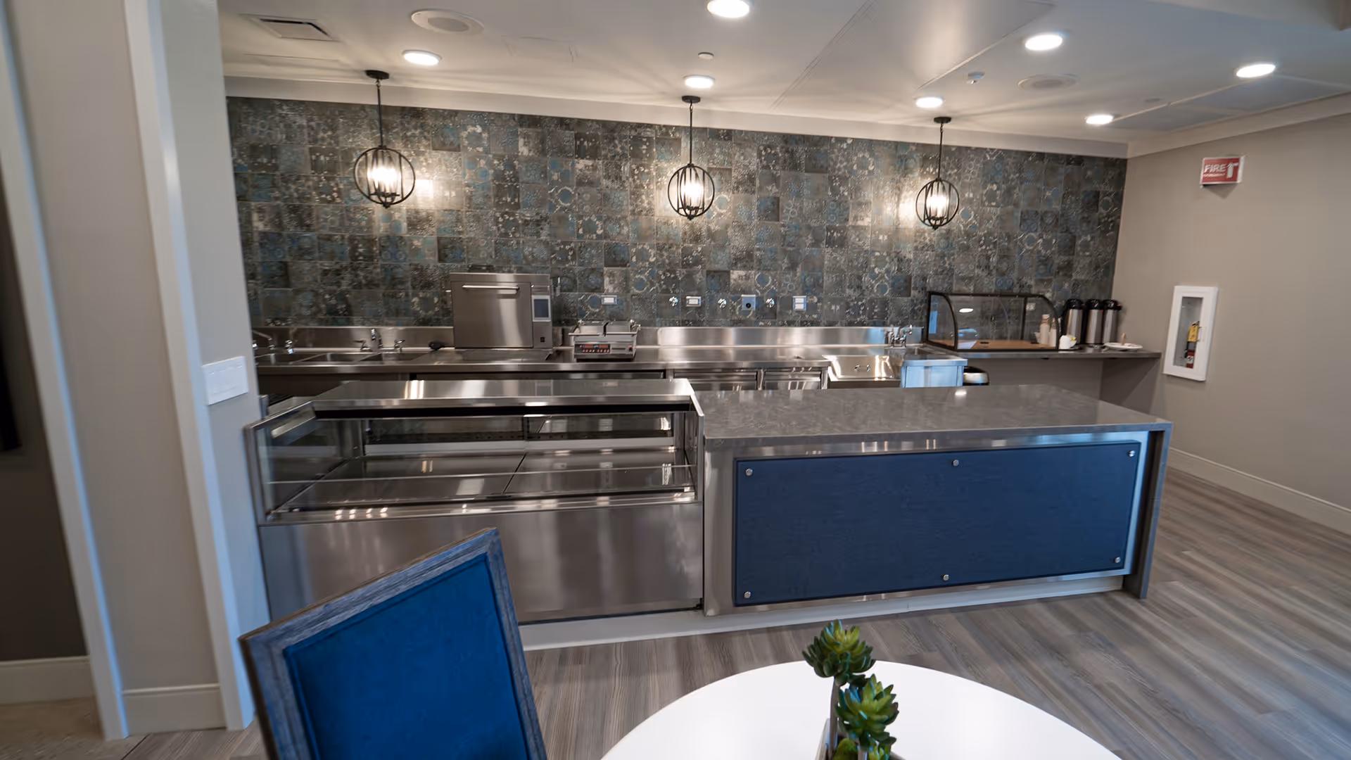 Modern kitchen area with stainless steel appliances and counters, a decorative tiled backsplash, three hanging pendant lights, and a small table with a blue chair and a plant in the foreground.